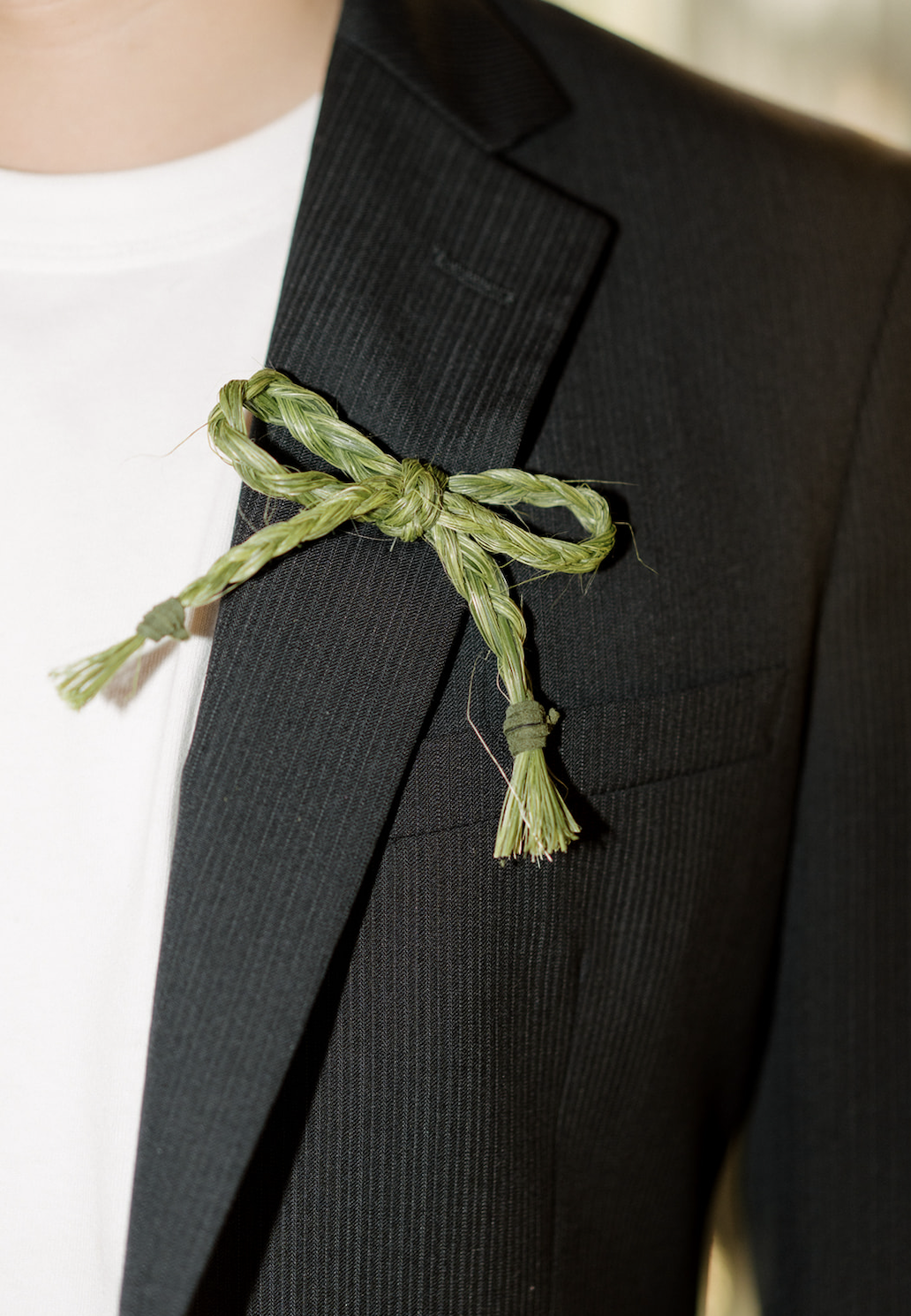 Close-up of a person wearing a black suit jacket with a white shirt underneath and a green braided rope tied as a decorative accessory on the lapel.