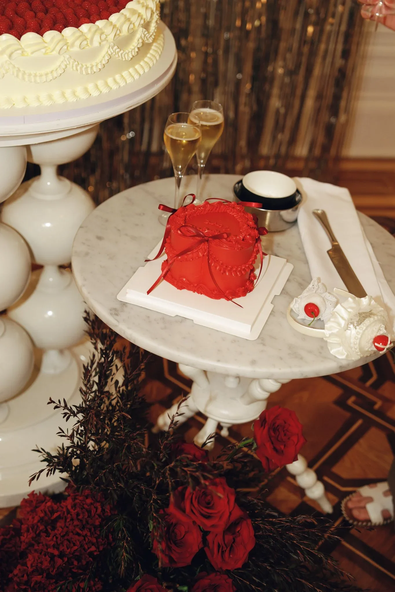 A table displays a red heart-shaped cake with chocolate icing and red ribbons, two glasses of champagne, and a small dish of whipped cream with cherries. In the background, there's a large round cake with white frosting and strawberries, and floral d