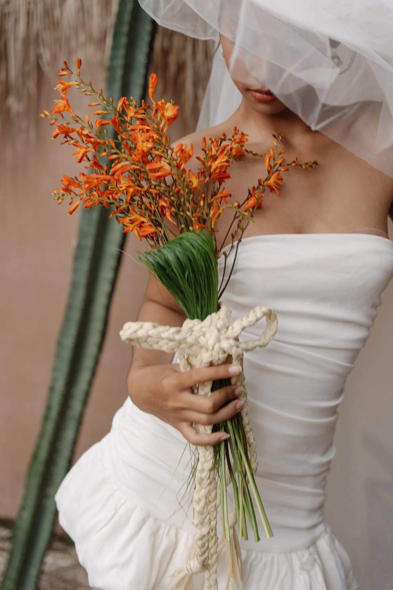 A woman wearing a white dress and veil holding a bouquet of orange flowers wrapped with a white braided rope, with a blurred outdoor background.
