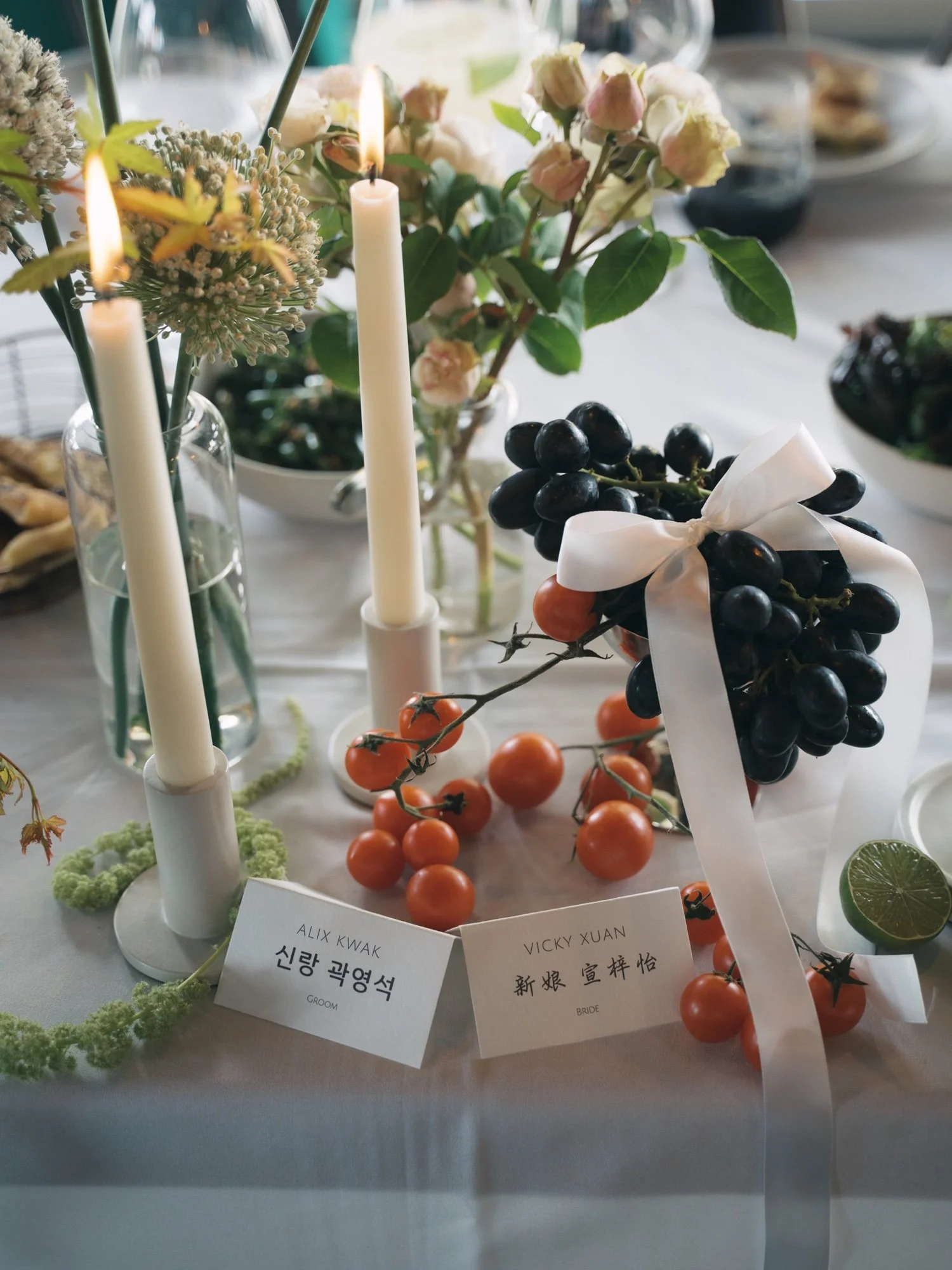 Various flowers, grapes with white ribbon, small orange tomatoes, and candles on a table, with name tags reading 'Groom' and 'Bride'.