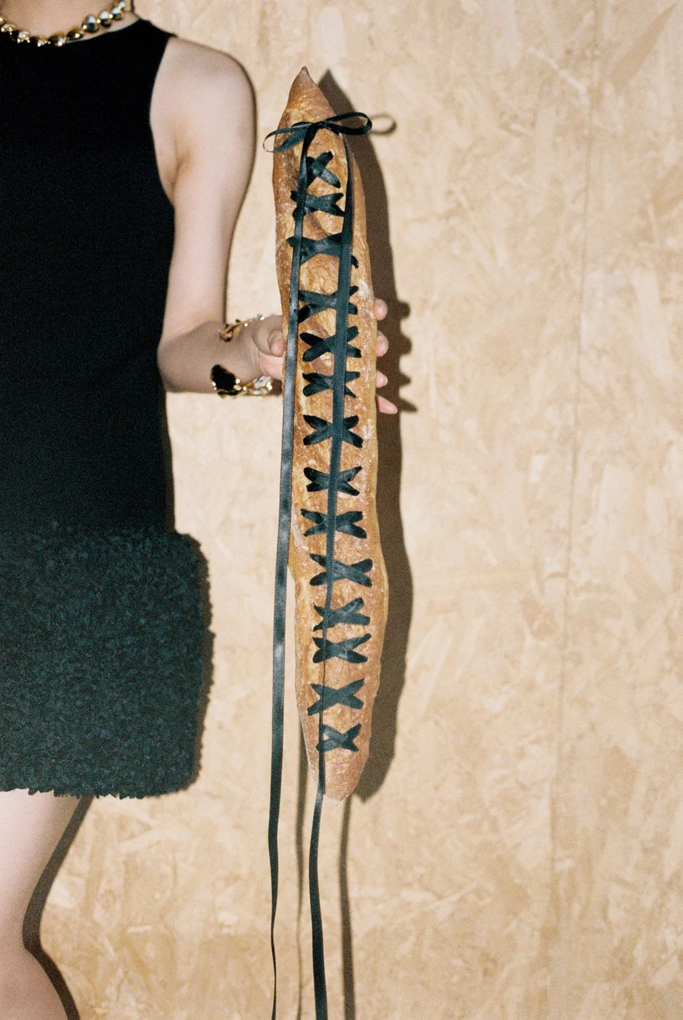 A person holding a wooden bow with black decorative material laced along its length. The person is dressed in a black outfit with gold jewelry and is standing against a beige wall.