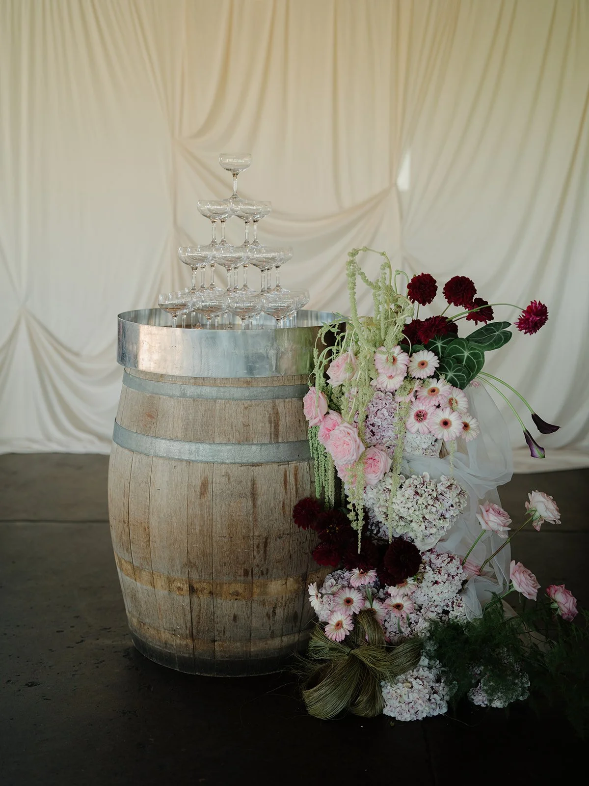 A wedding or celebration scene with a wooden barrel, champagne glasses stacked in a pyramid, and a large floral arrangement with pink, white, and dark red flowers, set against a cream-colored draped fabric background.