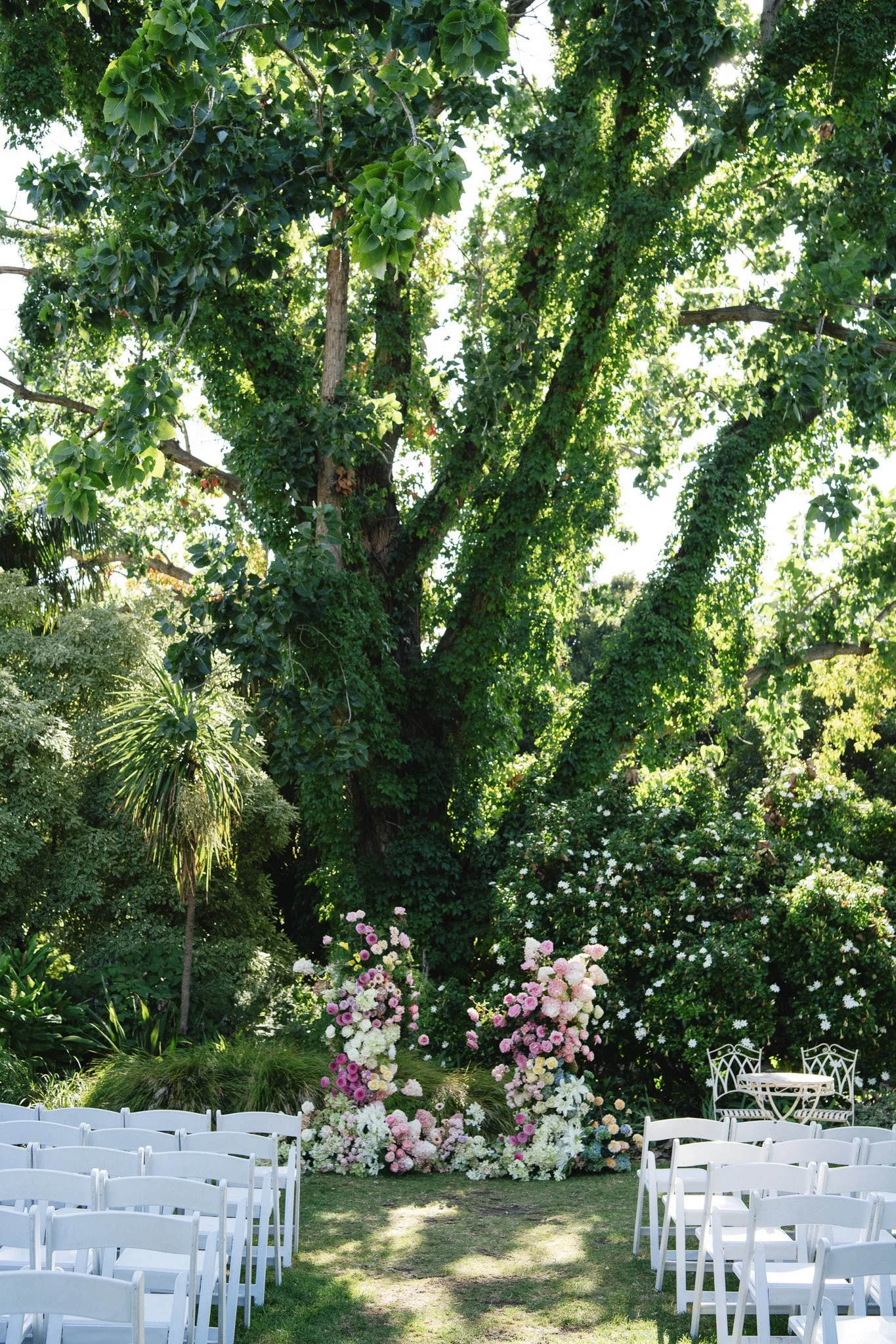 Outdoor wedding ceremony setup in a garden, with white chairs arranged on grass facing a floral arch with pink and white flowers, surrounded by lush green trees and bushes.