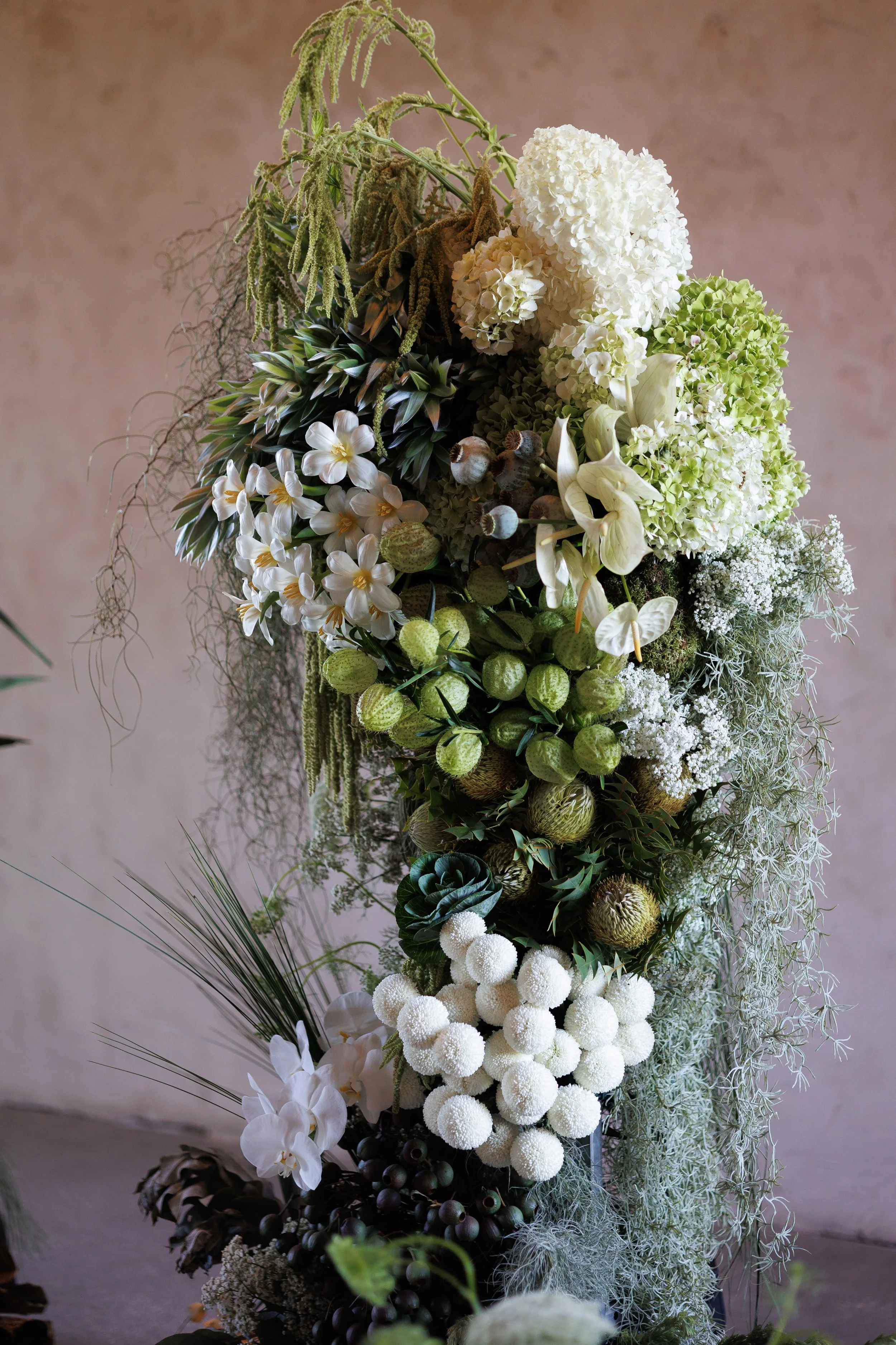 A tall floral arrangement with white hydrangeas, white orchids, green berries, and various greenery against a plain background.