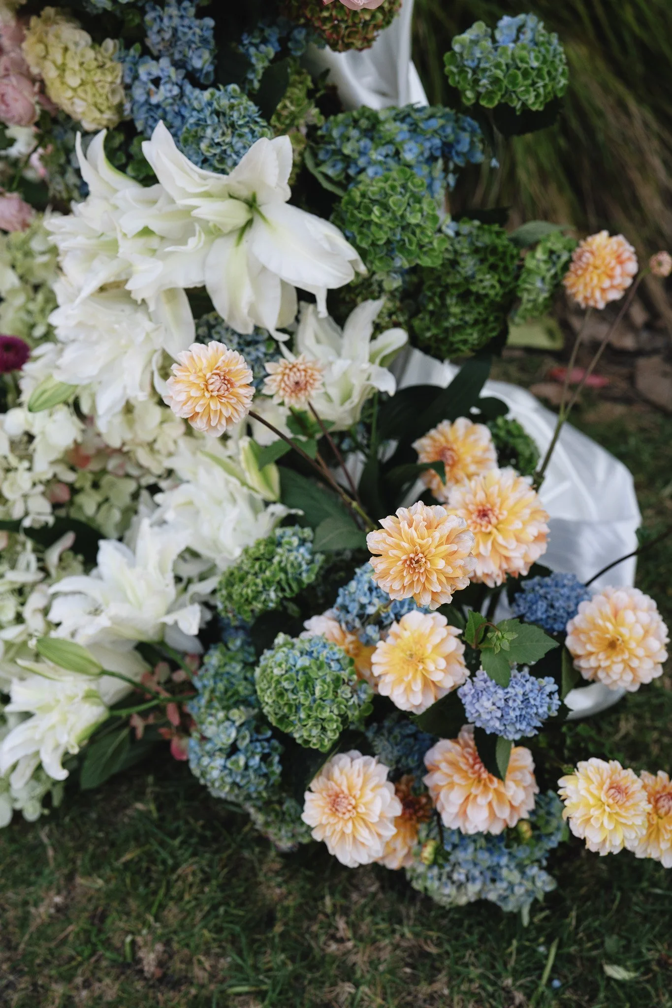 A close-up view of a colorful assortment of flowers, including white lilies, peach dahlias, blue hydrangeas, and green foliage, arranged outdoors on grass.