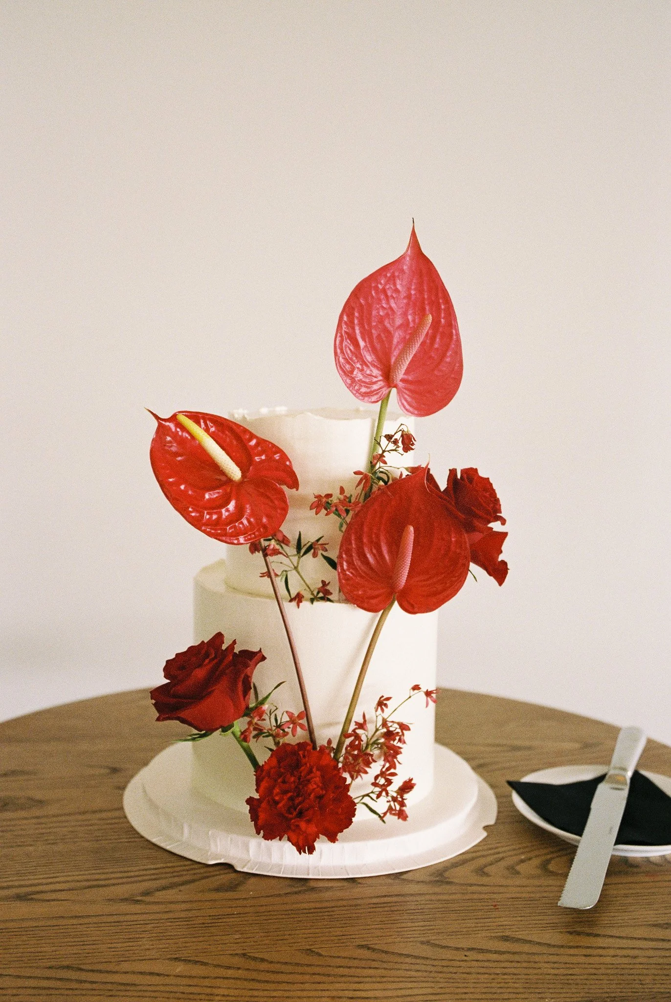 A white cake with red anthurium flowers and red carnations on it, placed on a white paper board on a wooden table, with a black napkin and knife beside it.