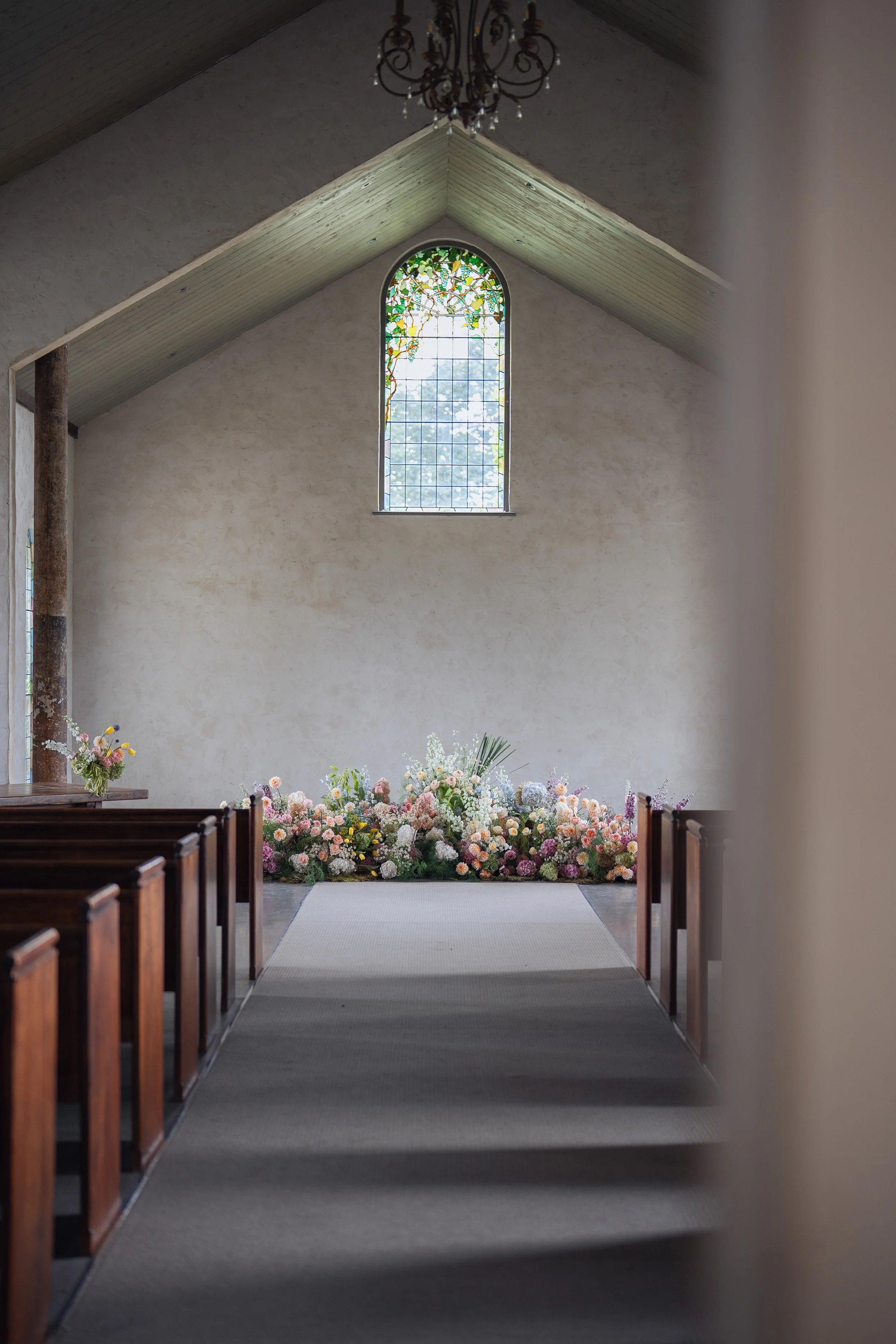 Inside a small church with a high, vaulted ceiling and a large stained glass window behind the altar, decorated with numerous pink, white, and purple flowers.