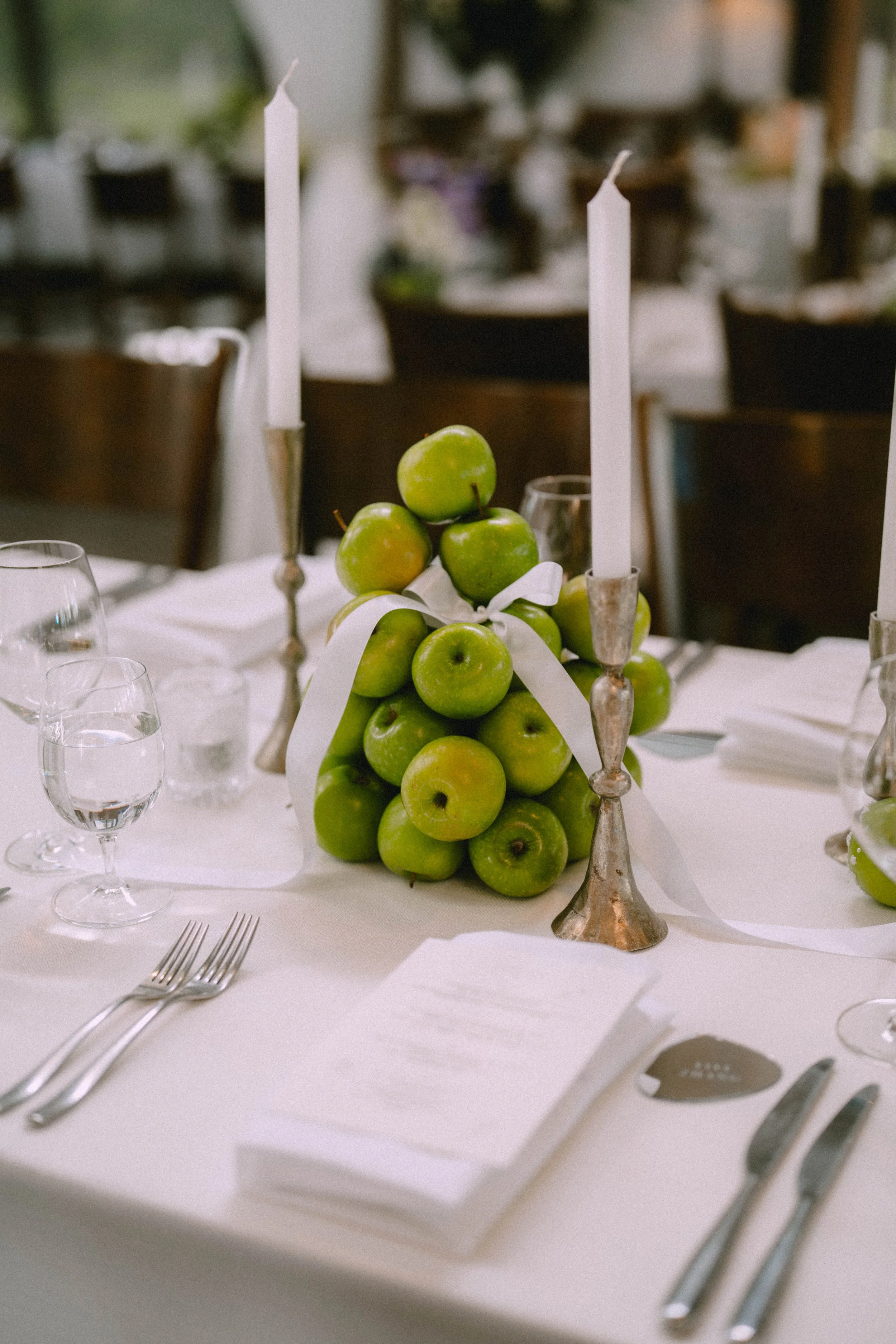 A formal table setting with a centerpiece of green apples tied with a white ribbon, surrounded by white candles in silver holders, and neatly arranged cutlery and glasses on a white tablecloth.