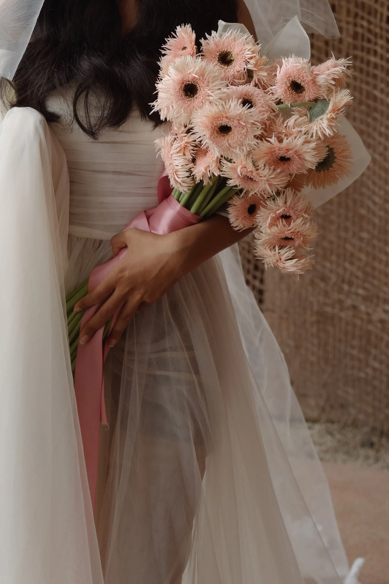 A woman in a wedding dress holding a bouquet of pink gerbera daisies wrapped in pink ribbon.