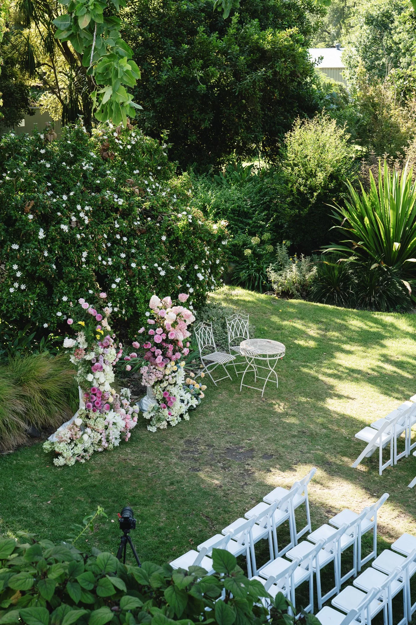 An outdoor wedding setup in a lush garden with white chairs arranged in rows and a floral arch with pink and white flowers. There is a small round table with chairs, surrounded by green plants and trees.