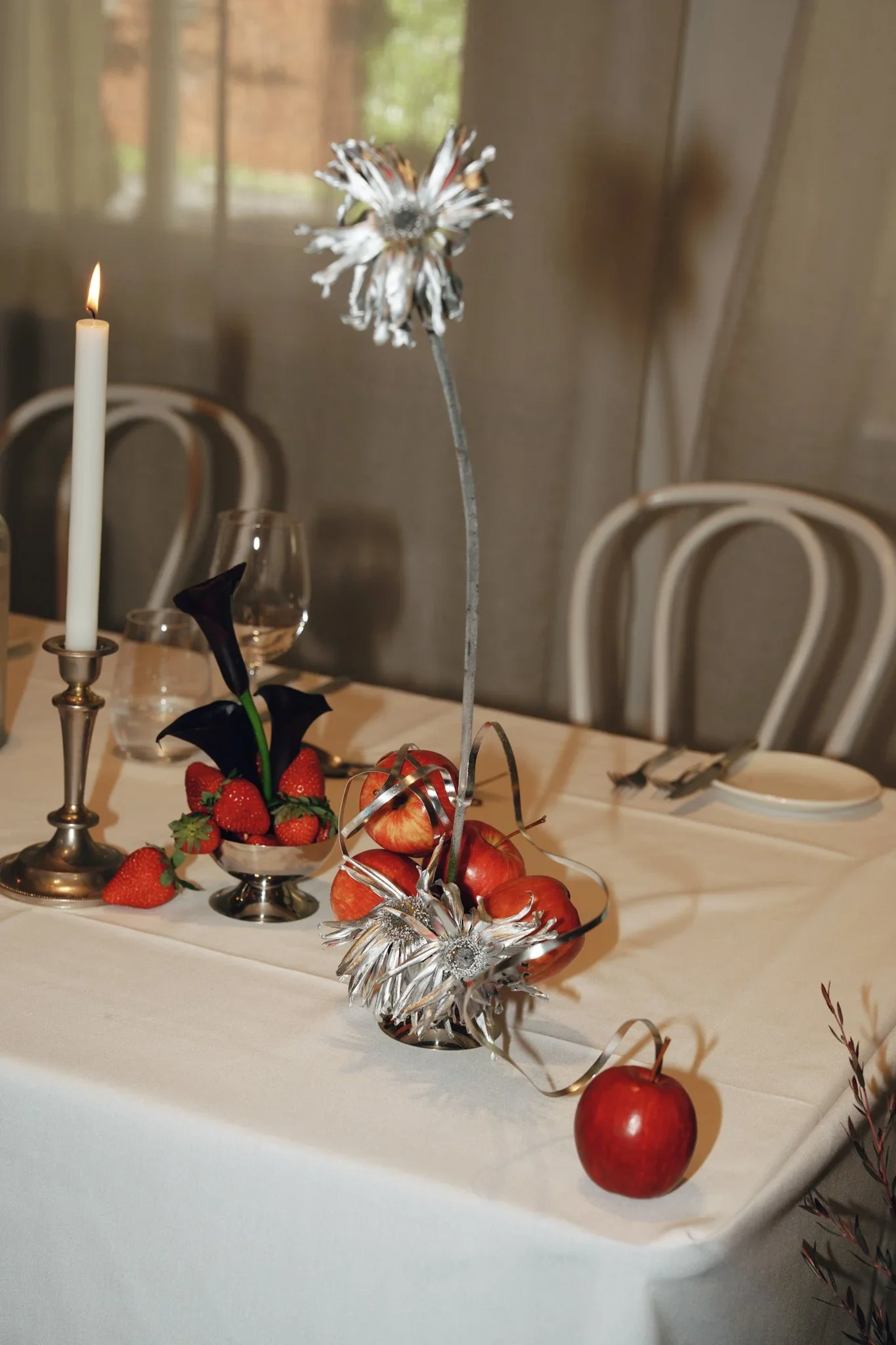 Table decorated with a silver flower centerpiece, strawberries, apples, a white candle, and black calla lilies, set for a celebration or event in a well-lit room with beige chairs and cream-colored curtains.
