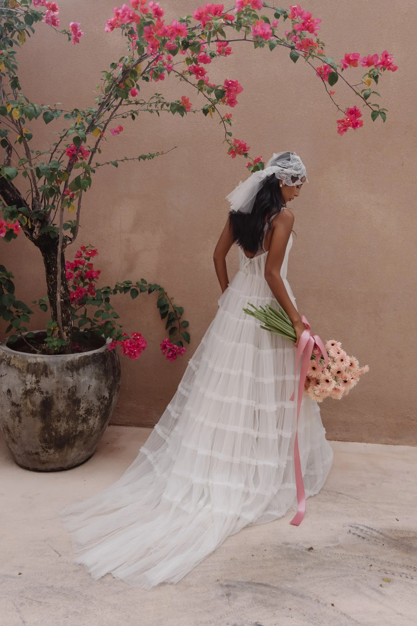 A woman in a flowing white wedding dress holding a large bouquet of pink (possibly blush) flowers with a pink ribbon, standing against a pastel pink wall. She is wearing a lace headpiece with a veil and has long dark hair. There is a large round pot 