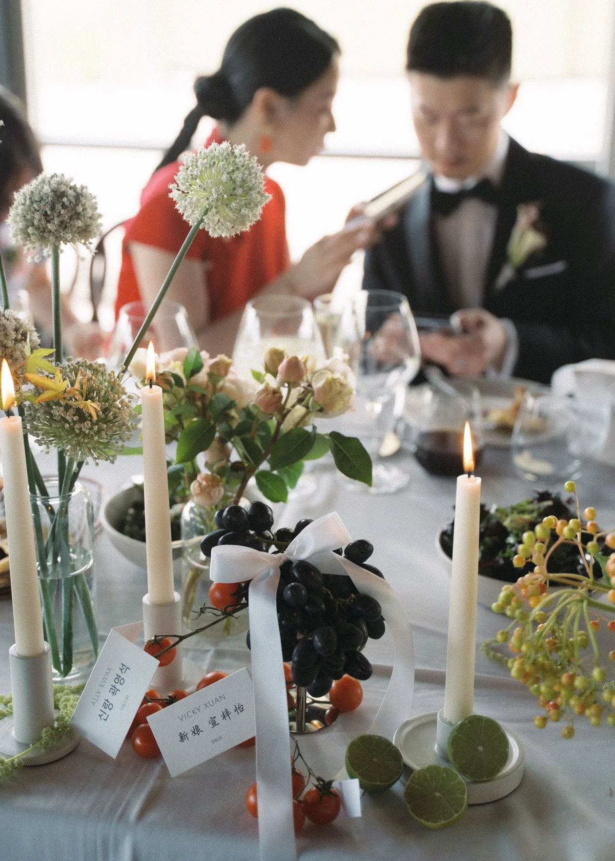 A wedding reception table decorated with flowers, candles, and fruit, with a bride and groom in the background.