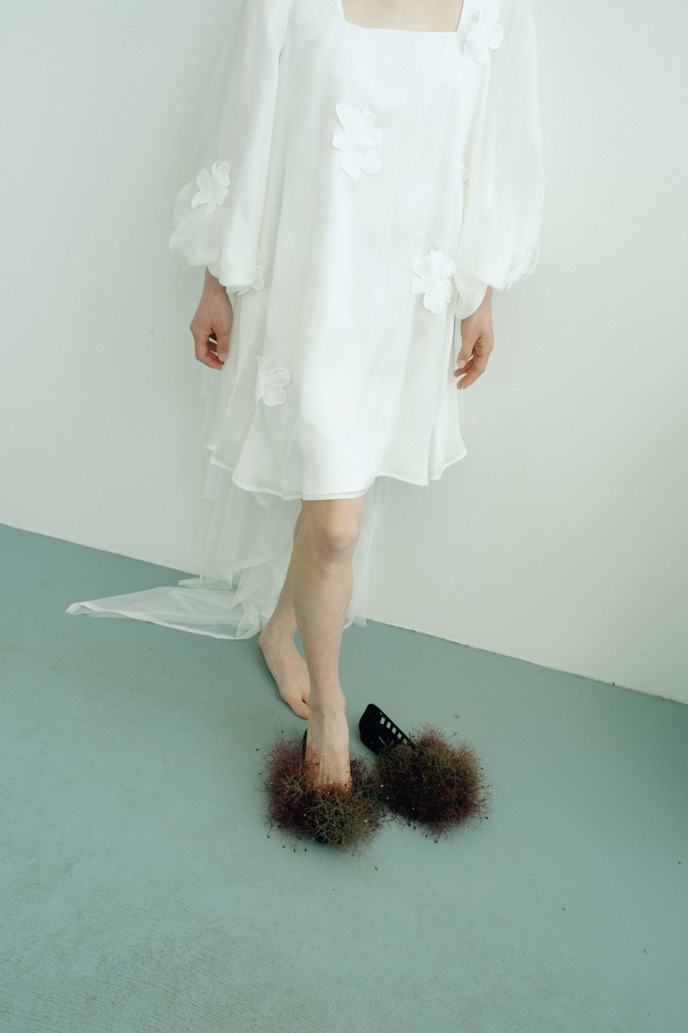 A mannequin or sculpture dressed in a white dress standing against a plain wall, with the lower half of the body replaced by a foot extending into a base of moss or grass.