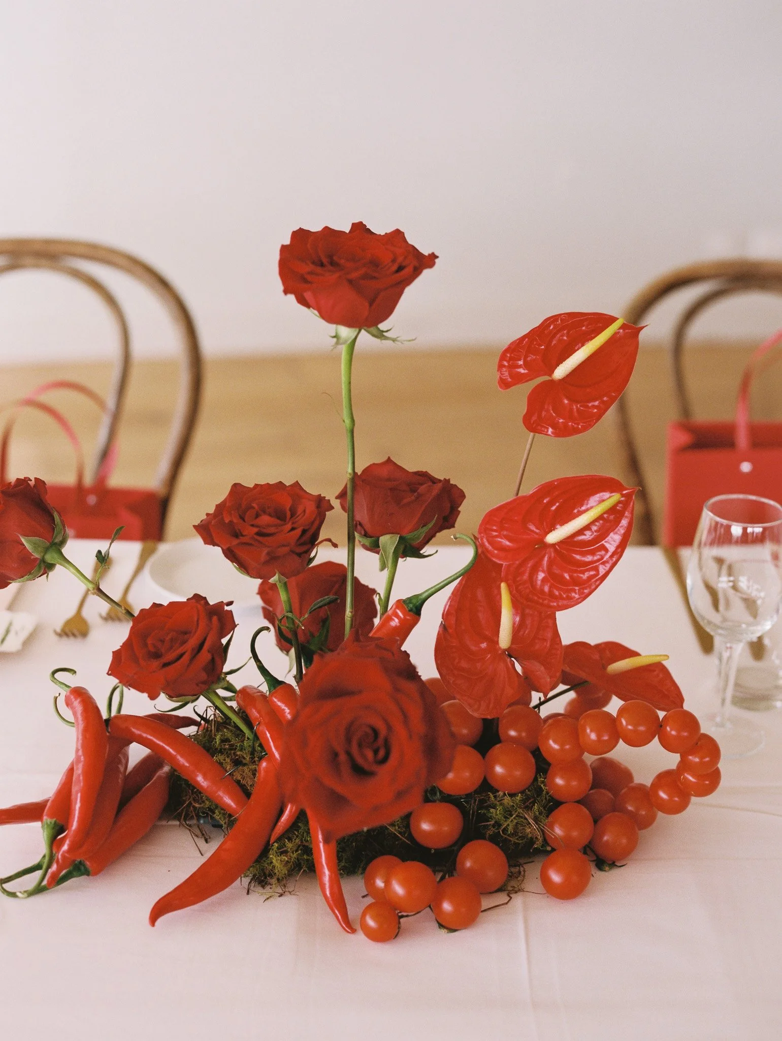 Arrangement of red roses, red anthuriums, red chili peppers, and orange beads on a white table.