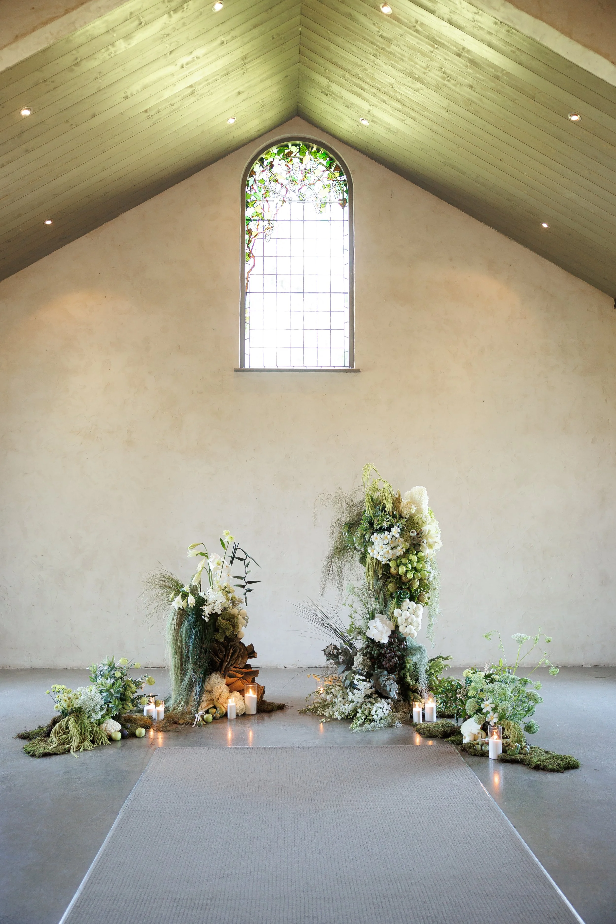 Interior of a church with a sloped wooden ceiling, stained glass window, and floral arrangements with white flowers and candles at the front.