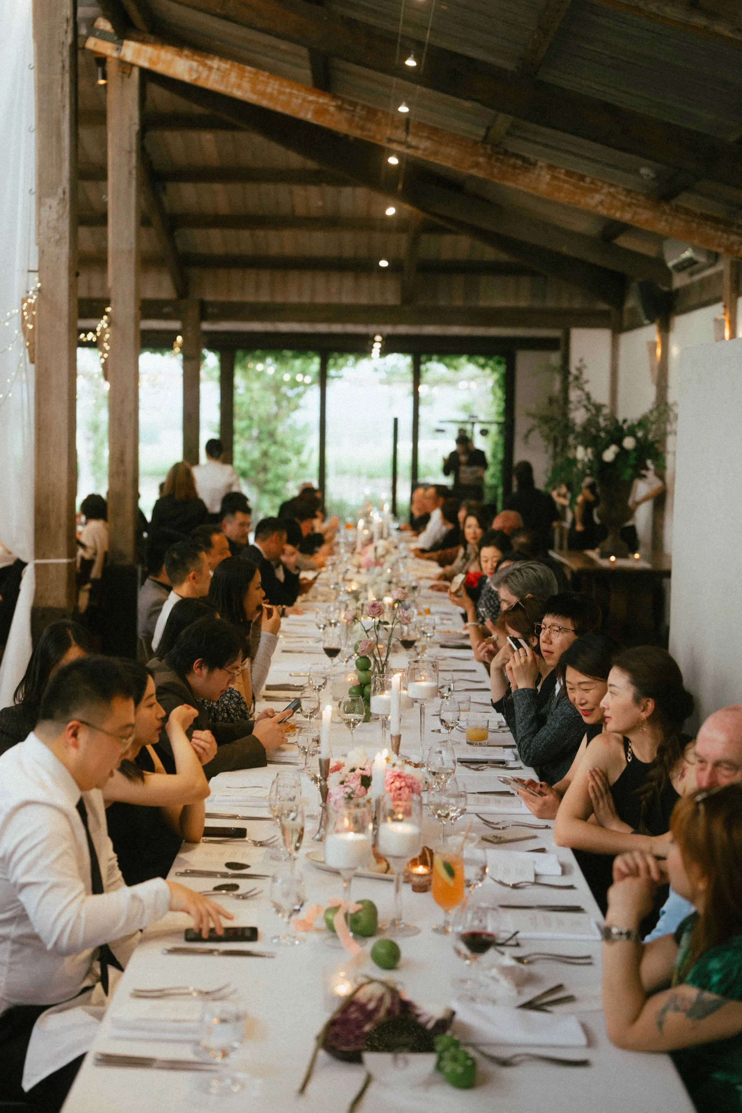 People sitting at a long dinner table inside a rustic venue with a high wooden ceiling, decorated with flowers and candles, during a social event or celebration.