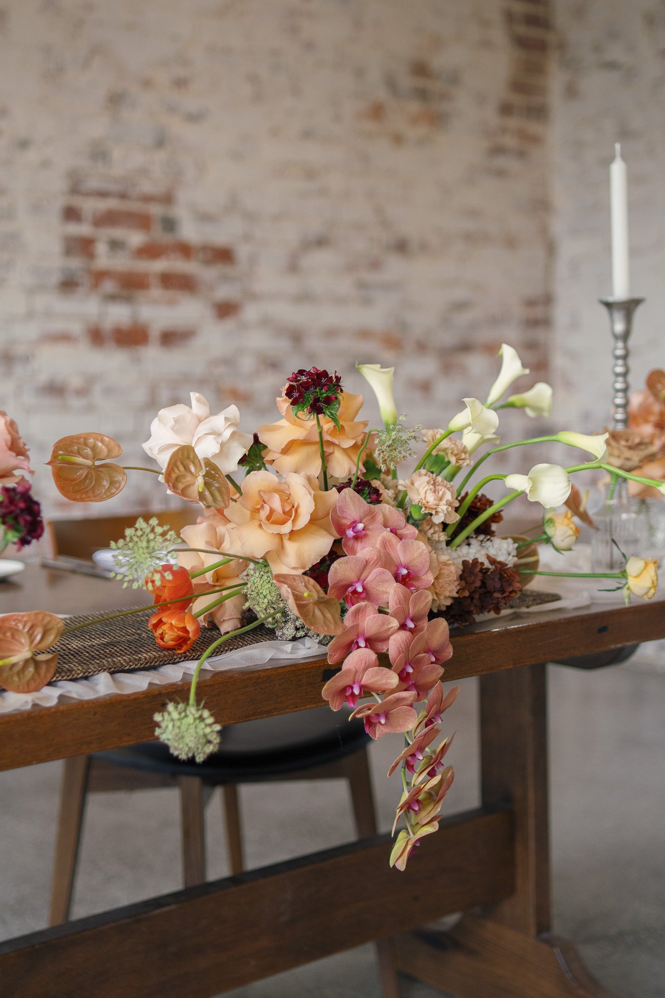 A floral arrangement on a wooden table with a stone brick wall in the background, featuring white calla lilies, peach, pink, and burgundy flowers, and a tall white candle in a silver candle holder to the right.