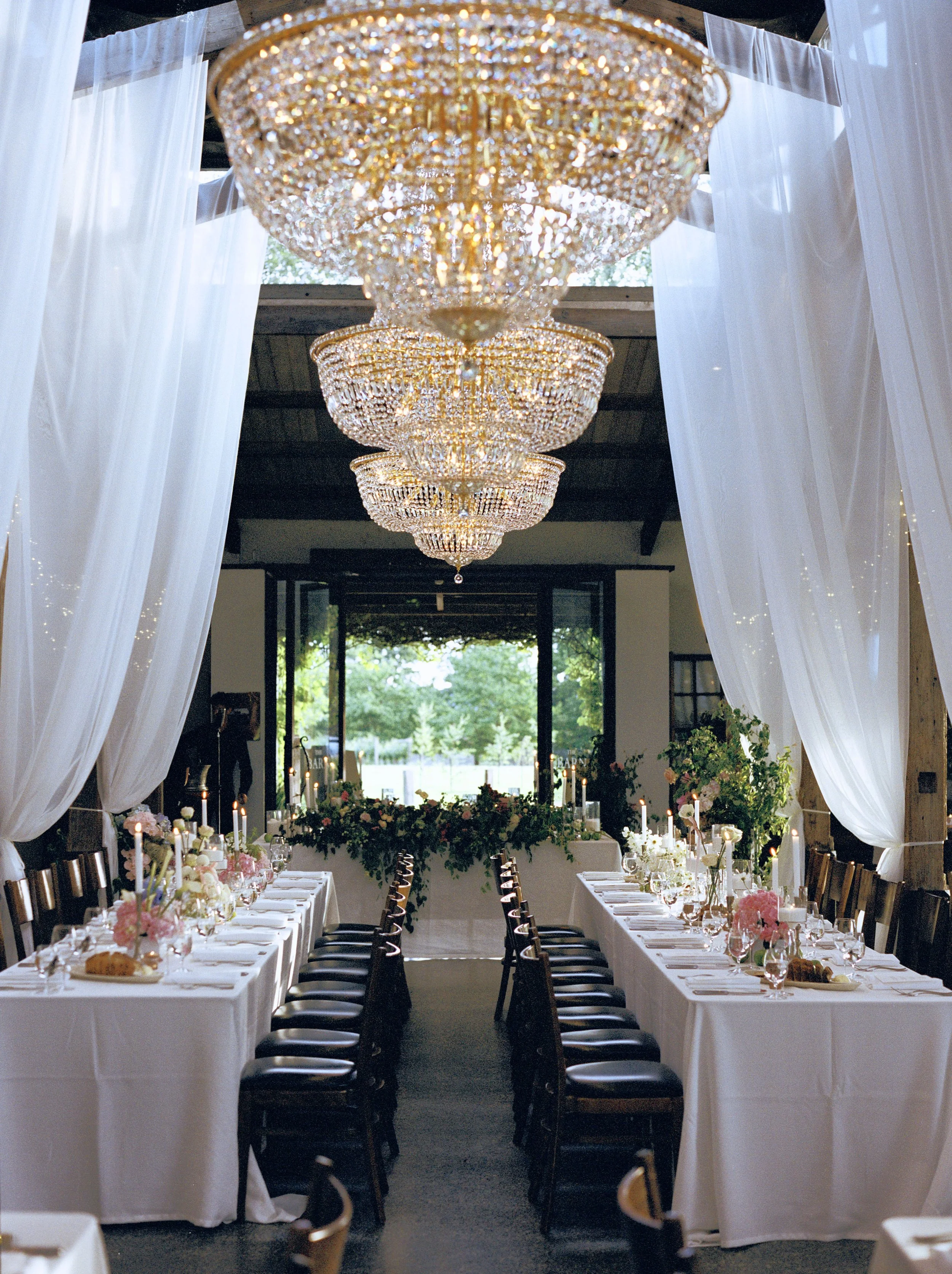 Elegant dining setup with long tables covered in white tablecloths, pink floral centerpieces, candles, and chairs arranged on either side. Large, ornate crystal chandeliers hang from the ceiling, and sheer white curtains frame the space, creating a l