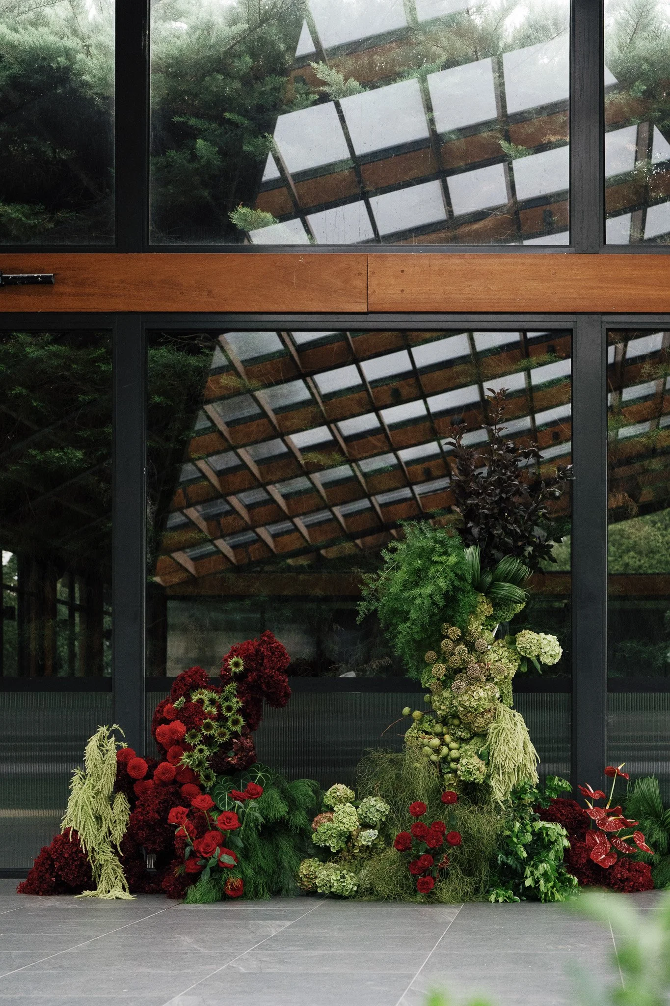A variety of vibrant red, green, white, and cream floral arrangements and foliage set against large glass windows showing a roof structure and trees outside.