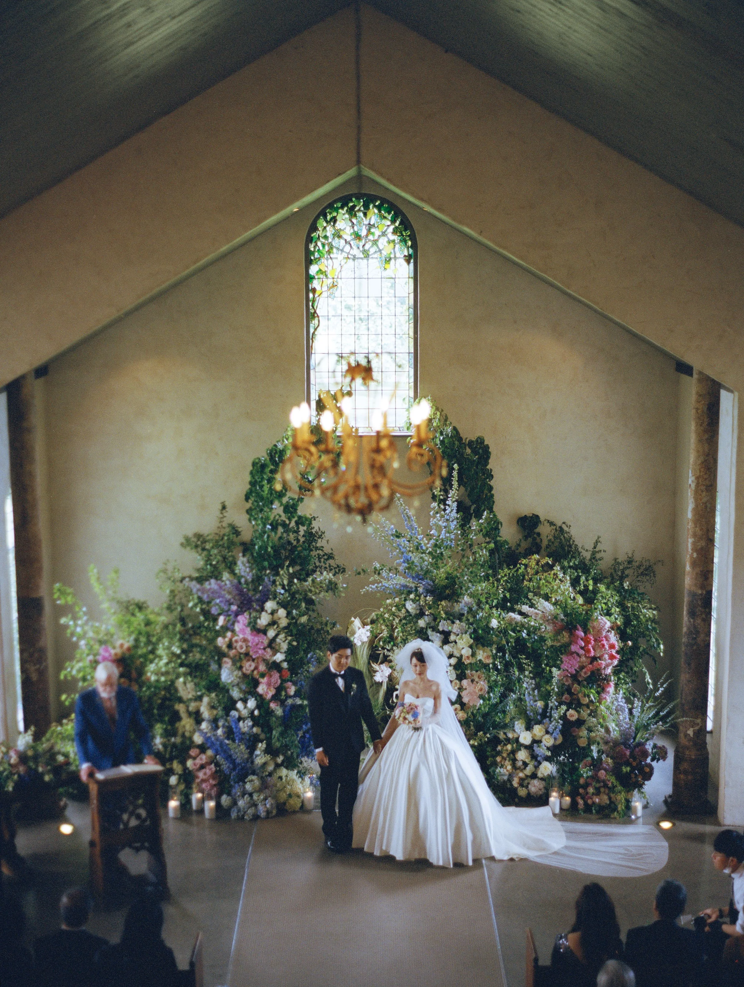 A wedding ceremony inside a church with a bride and groom standing at the altar, surrounded by large floral arrangements and greenery, with a stained glass window in the background and guests seated in pews.
