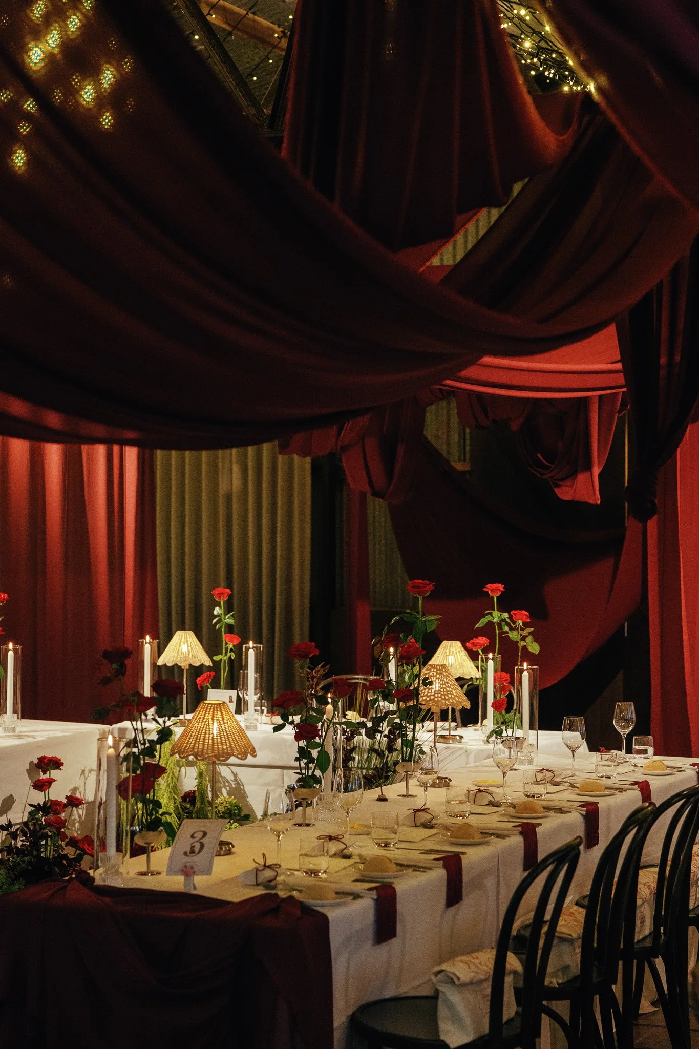 A decorated dining table with red flowers, candles, and small lamps in a cozy, dimly-lit restaurant or event space with wooden walls and curtains.