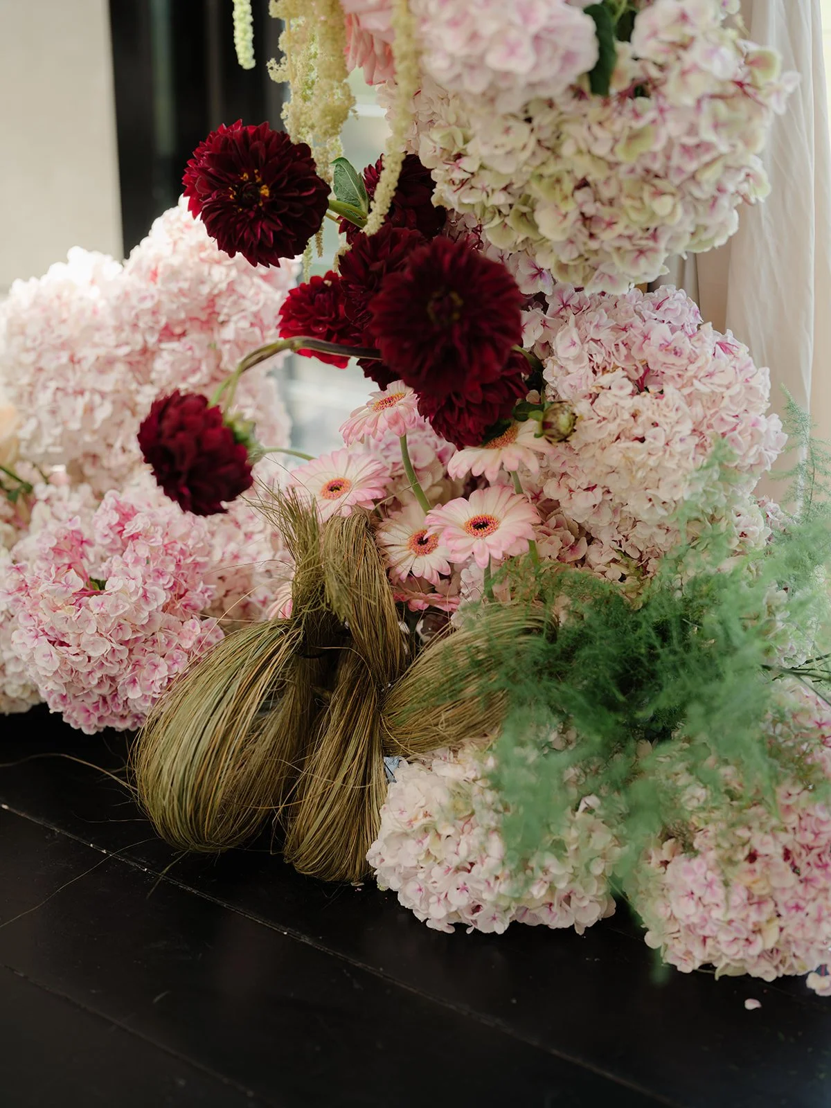 A bouquet of pink, white, and deep red flowers includes hydrangeas, gerbera daisies, and dahlias, arranged with natural greenery and decorative straw-like elements.