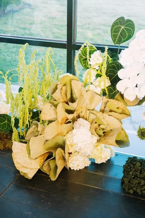 Arrangement of dried leaves and white flowers with green leafy plants in the background near a window.