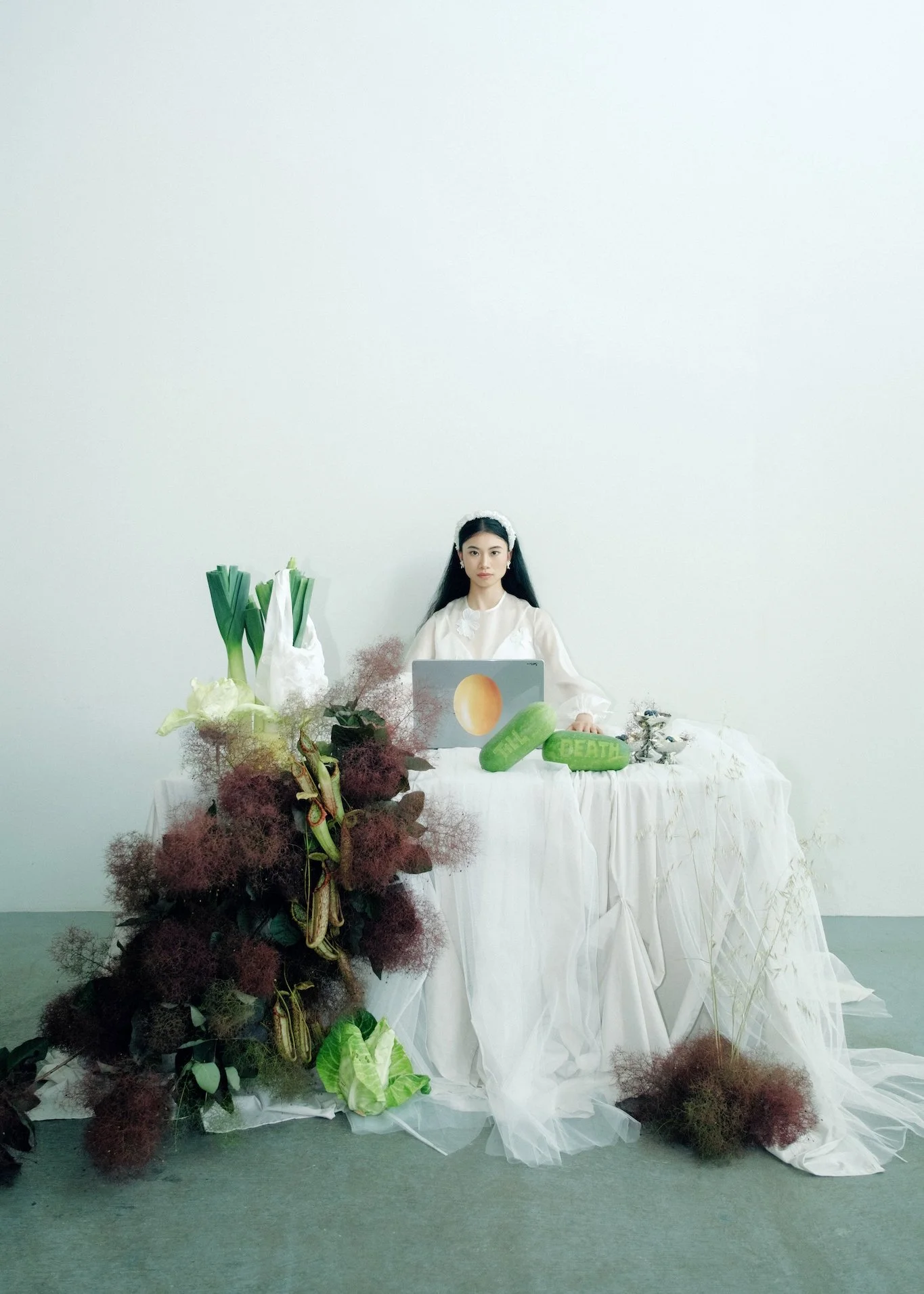 A woman sits behind a table covered with white cloth, surrounded by various plants and objects, with a plain white wall in the background.
