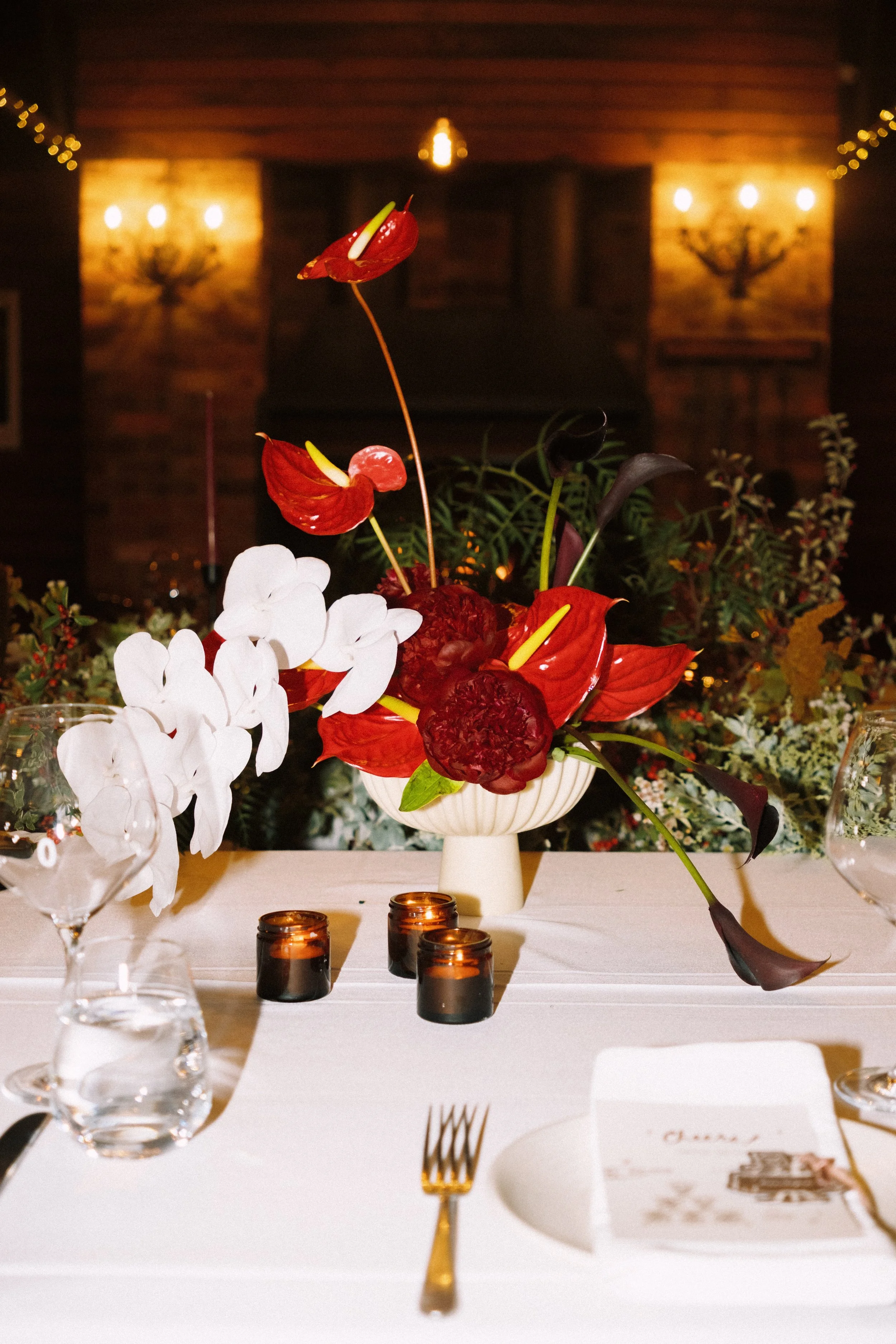 Elegant table setting featuring a white tablecloth, floral centerpiece with red and white flowers, surrounded by candles, wine glasses, and utensils.