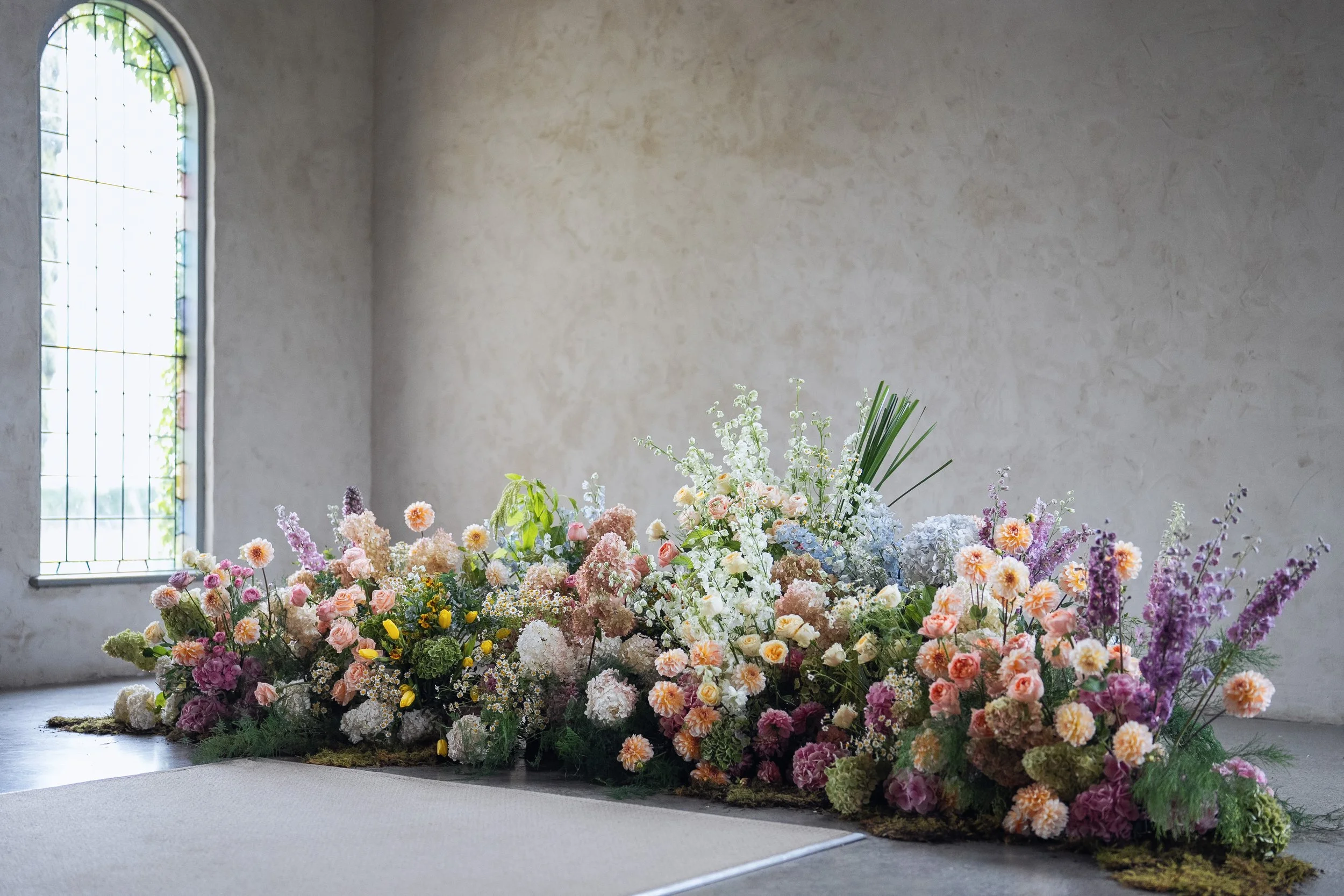A large, colorful floral arrangement placed on the floor in a decorated indoor space with a high, textured wall and a large arched window with stained glass.