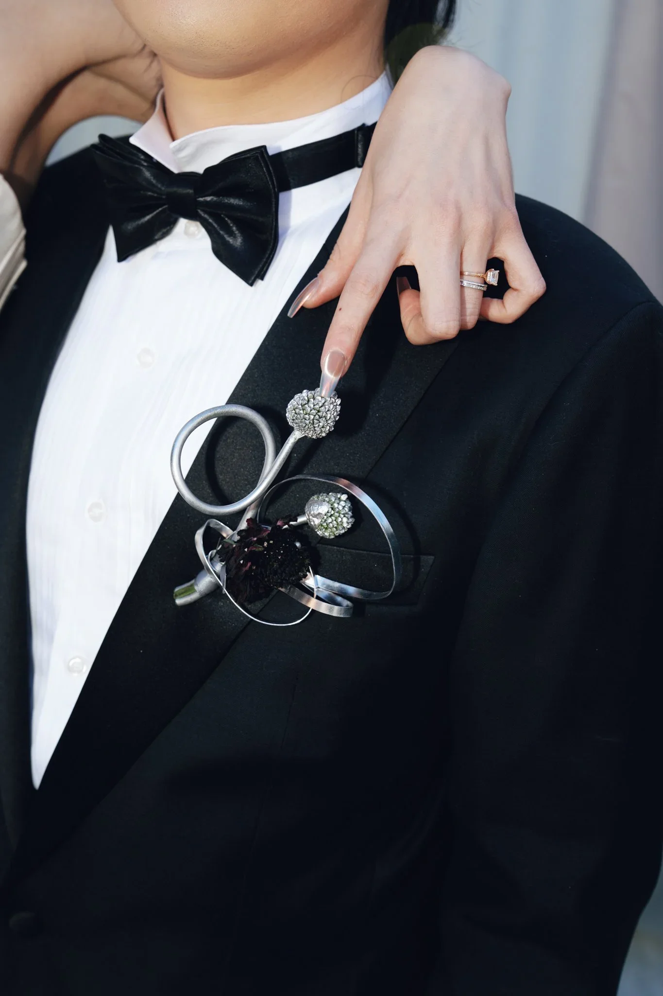 A person wearing a tuxedo with a bowtie, holding a spoon with a jeweled ring on their finger, and some jewelry on their lapel.