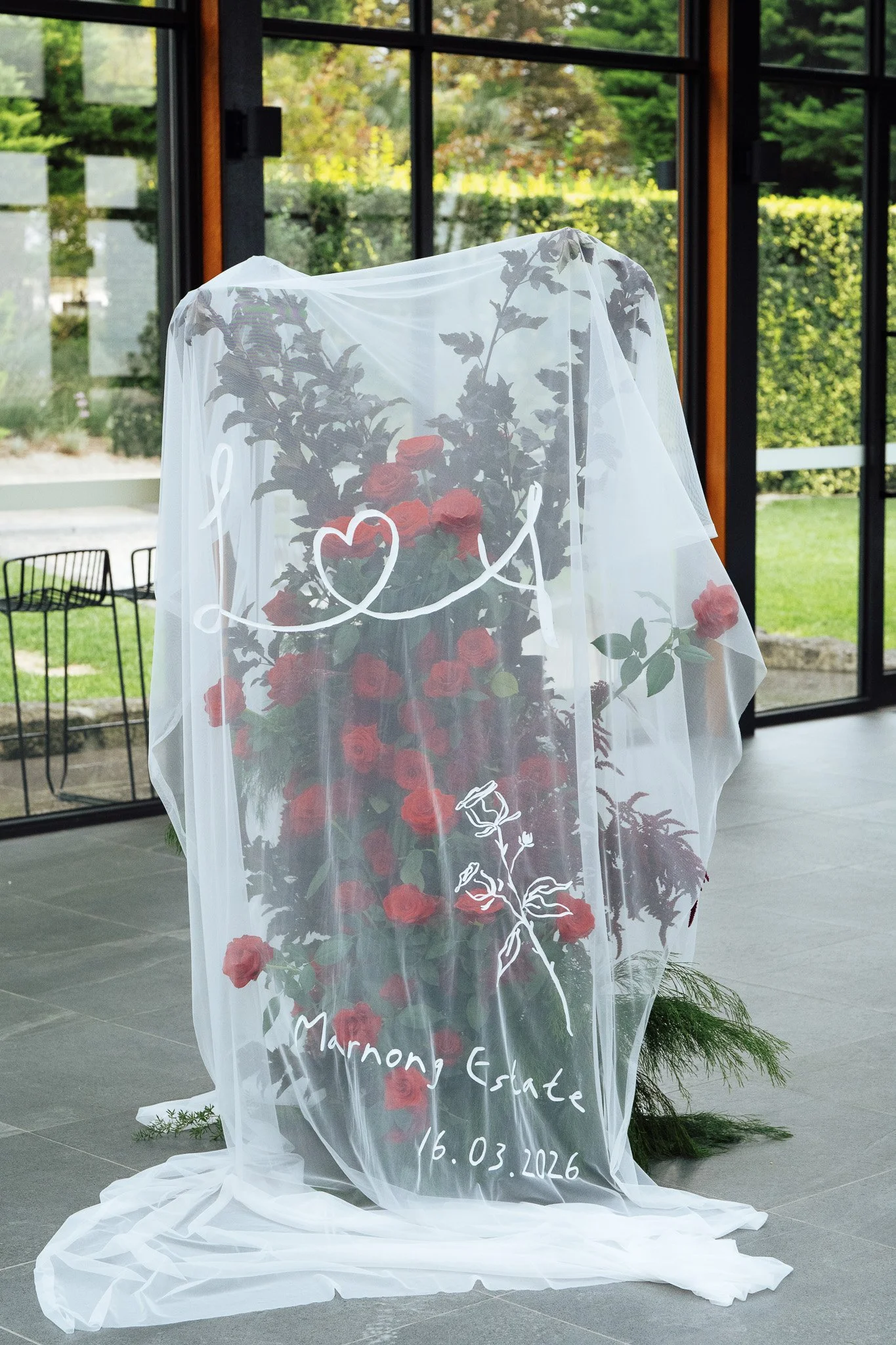 A floral arrangement of red roses is visible behind a white sheer curtain with the words 'Love Marriage Estate' and a date written in white. The curtain has decorative floral and heart designs, and the scene is set inside a room with large windows sh