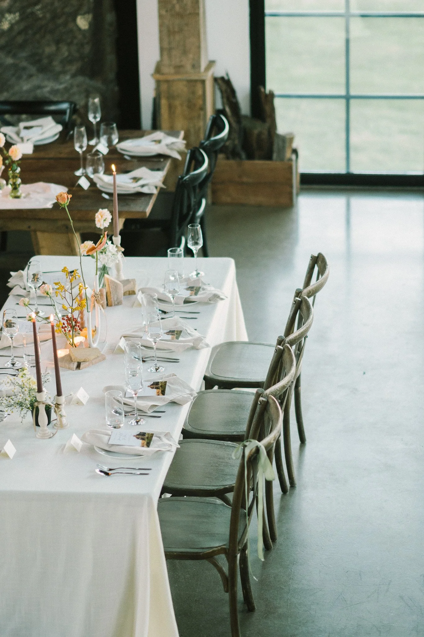 A decorated dining table set for a meal with white tablecloth, floral centerpieces, tall candles, and glassware in a modern, rustic-style room with wood chairs and a large window.