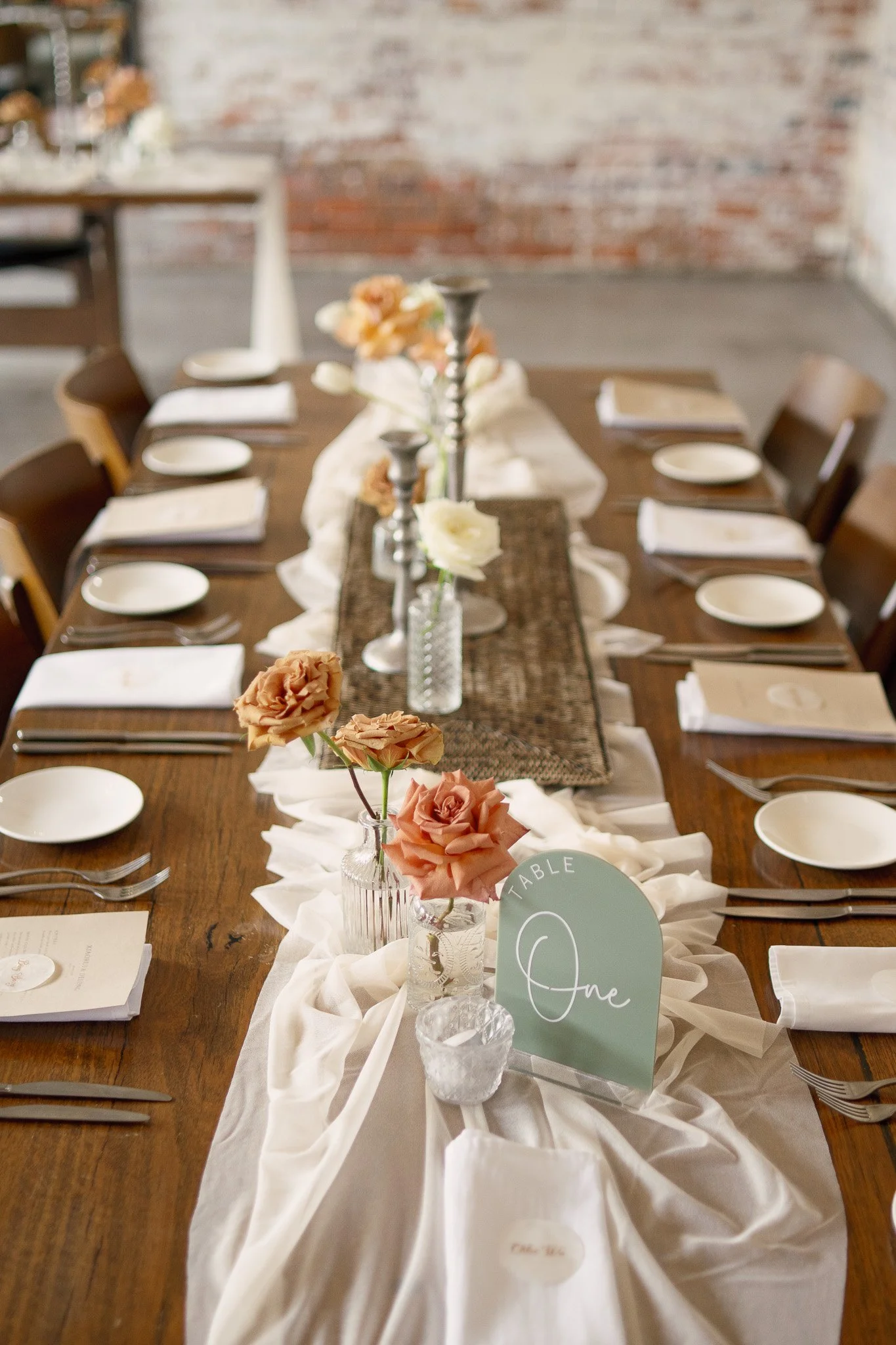 Table decorated for a special occasion with a white table runner, pink and cream roses in glass vases, place settings with white napkins, and a sign that reads 'Table 01 One' in a small glass holder.