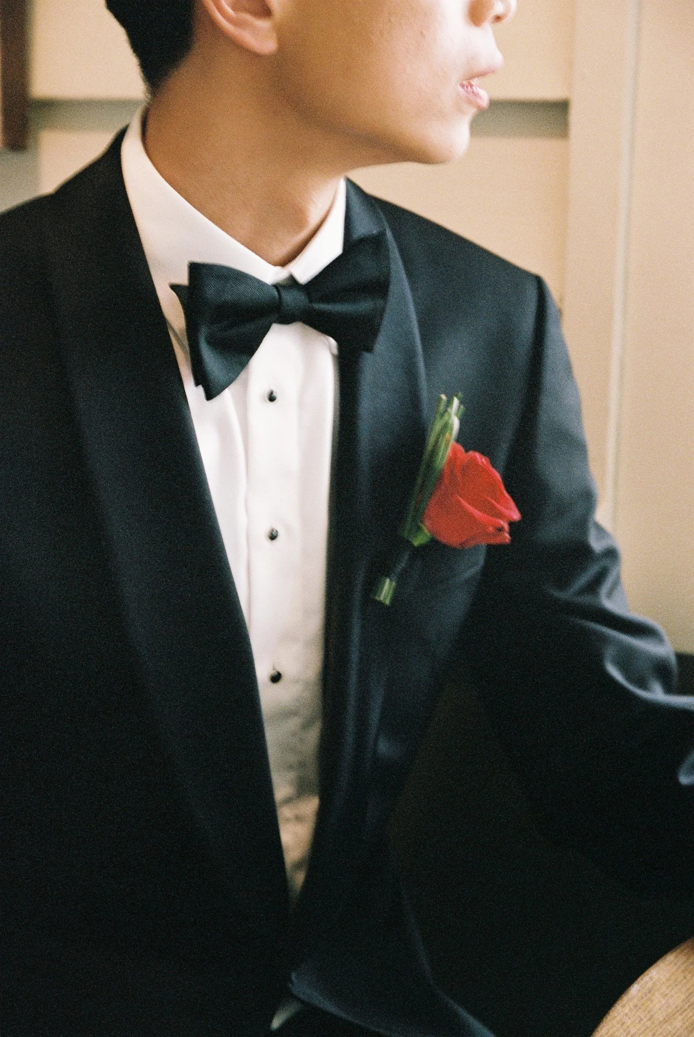 Close-up of a man in a tuxedo with a white shirt, black bow tie, and a red boutonniere, sitting in a formal setting.
