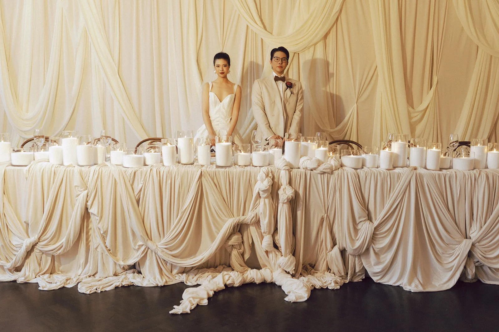 A bride and groom standing behind a decorated wedding reception table covered with cream-colored fabric and candles.