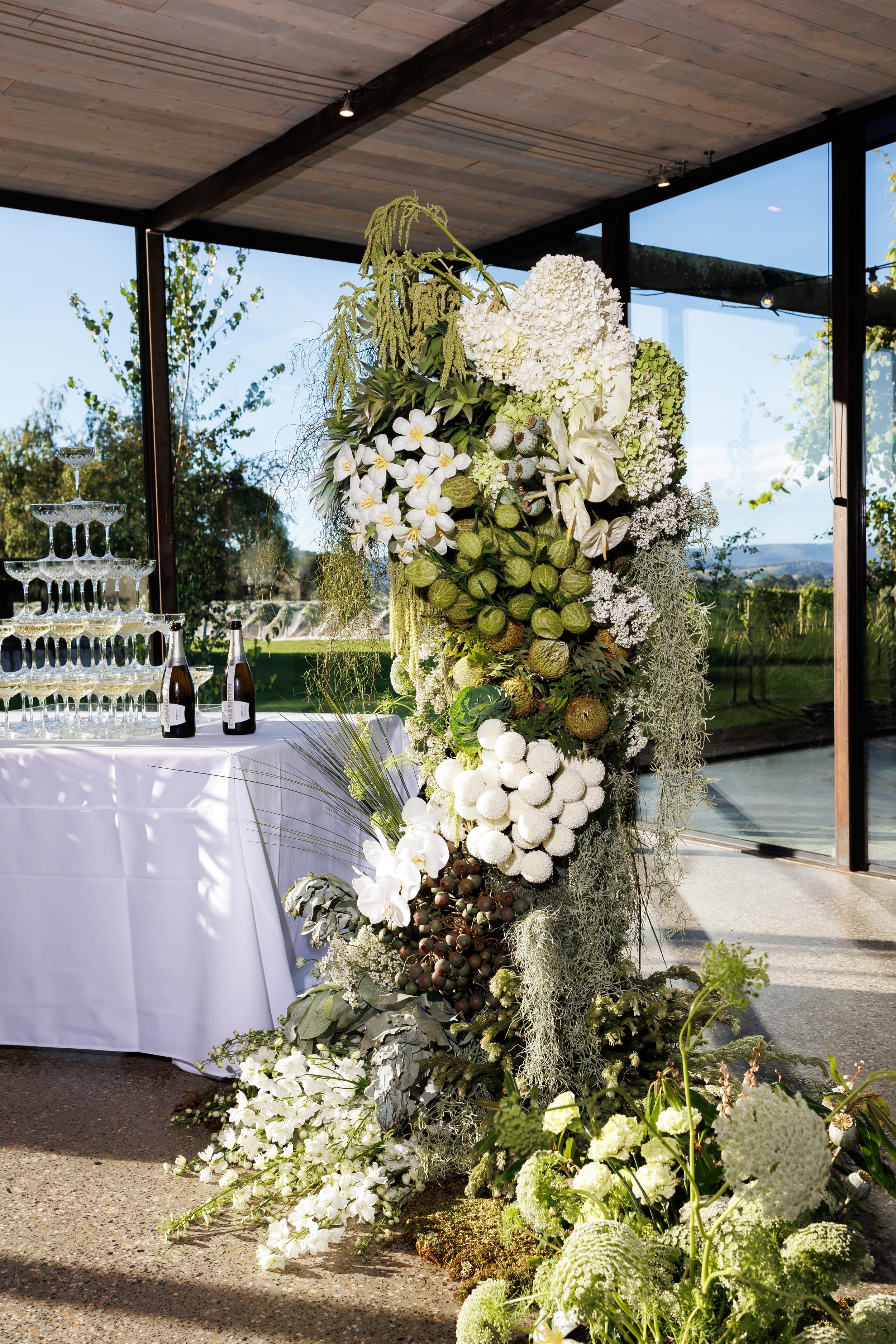 A tall floral arrangement of white and green flowers and foliage stands next to a table with a white tablecloth and champagne bottles. Glassware for champagne is stacked on the table. The setting appears to be indoors with large windows revealing a o