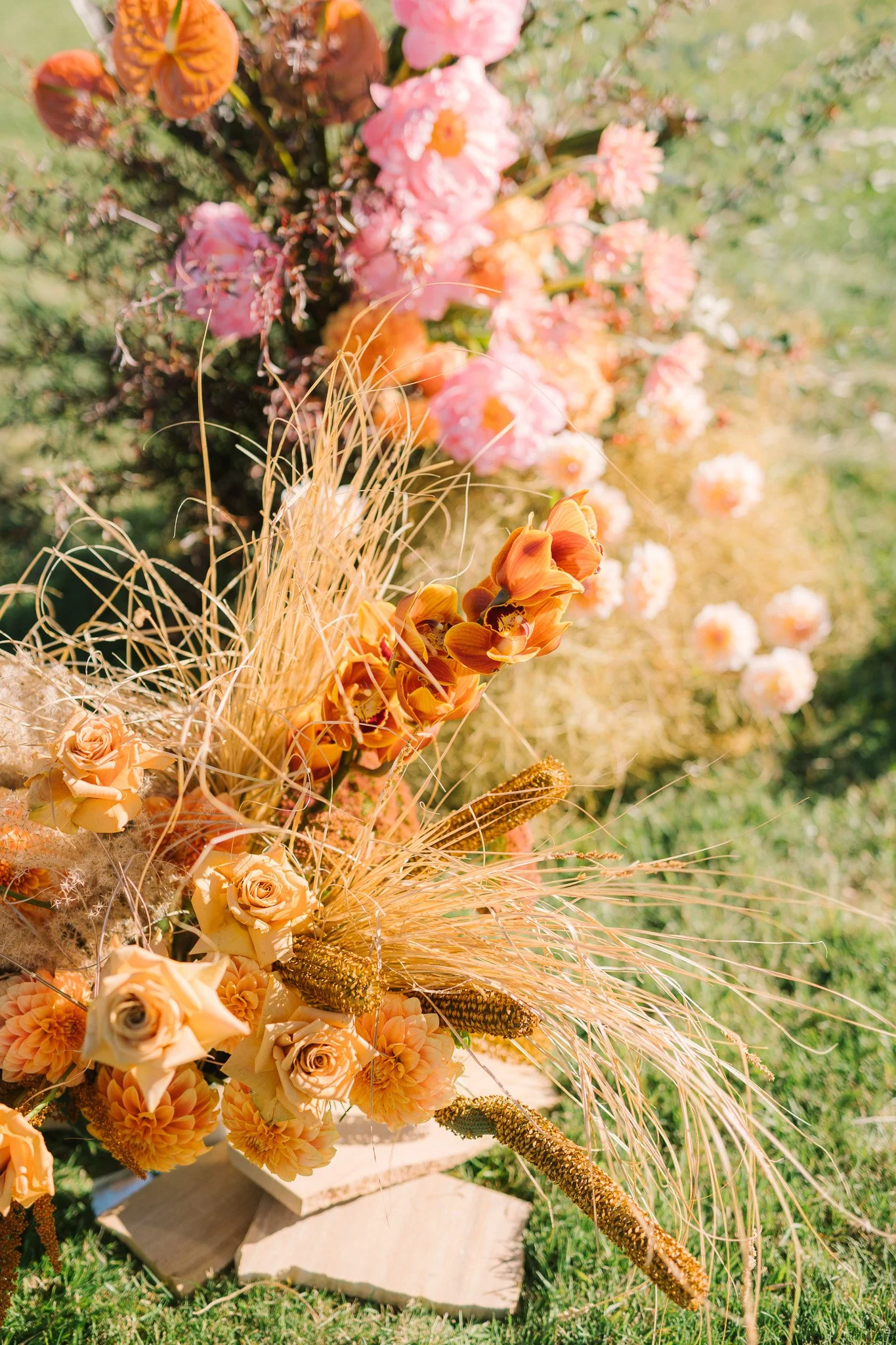 A flower arrangement with peach, pink, and orange flowers, accented by dry grasses and wheat stalks, placed on a white wooden stand outdoors on grass.