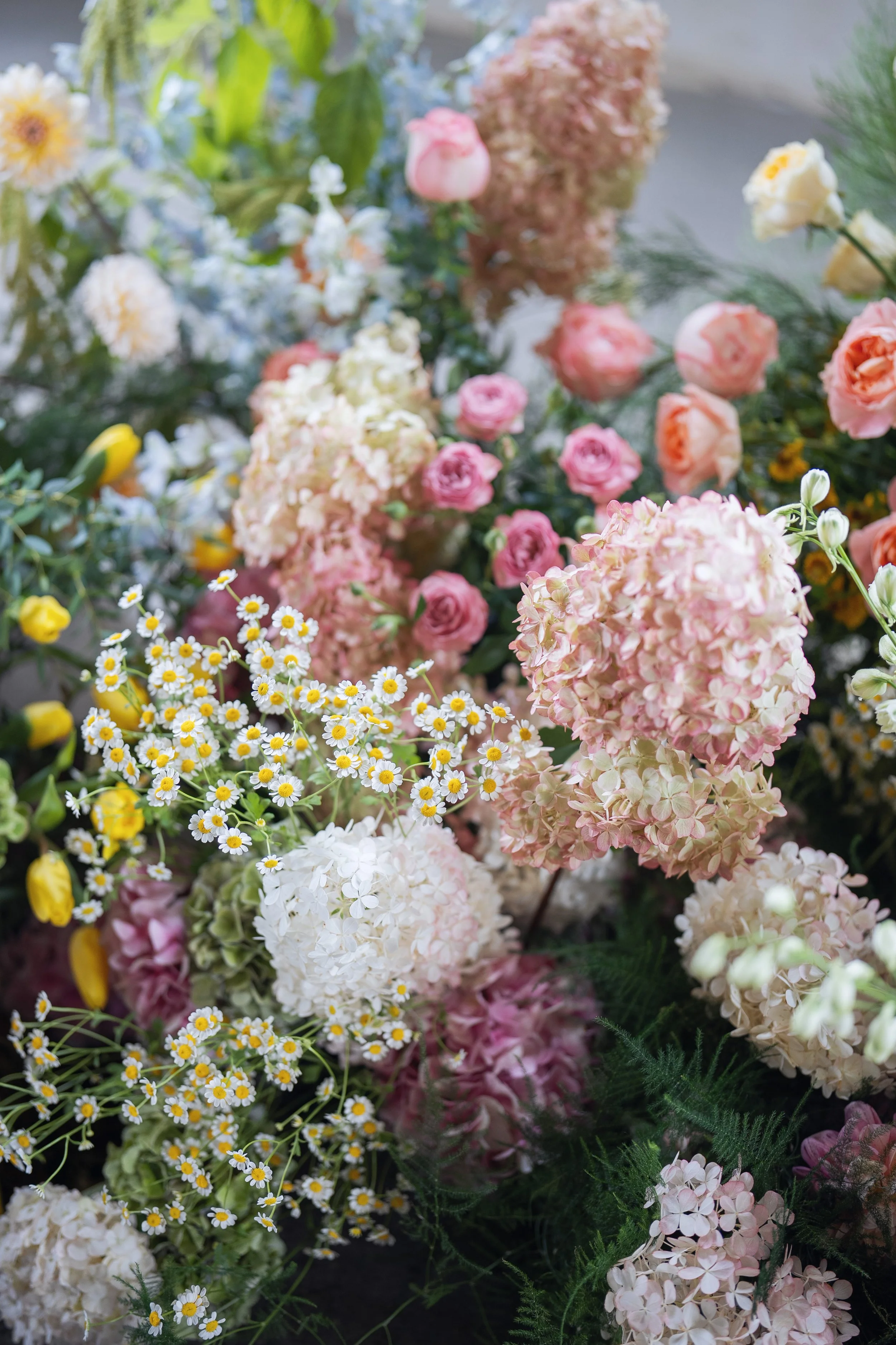 A close-up view of various colorful flowers, including pink and white hydrangeas, yellow daisies, and small white filler flowers, arranged in a floral display.