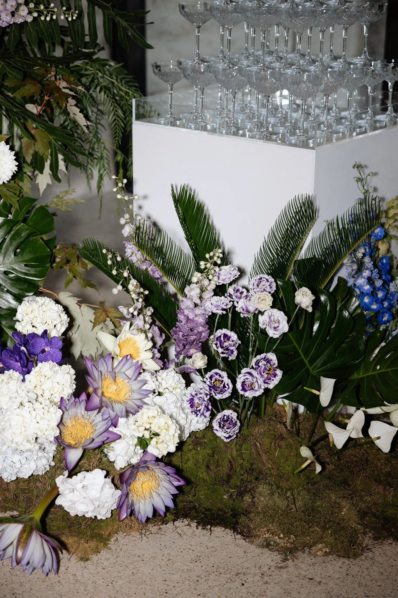 An arrangement of colorful flowers including white, purple, yellow, and blue blooms, with large green tropical leaves around them, situated outdoors on a sandy surface.