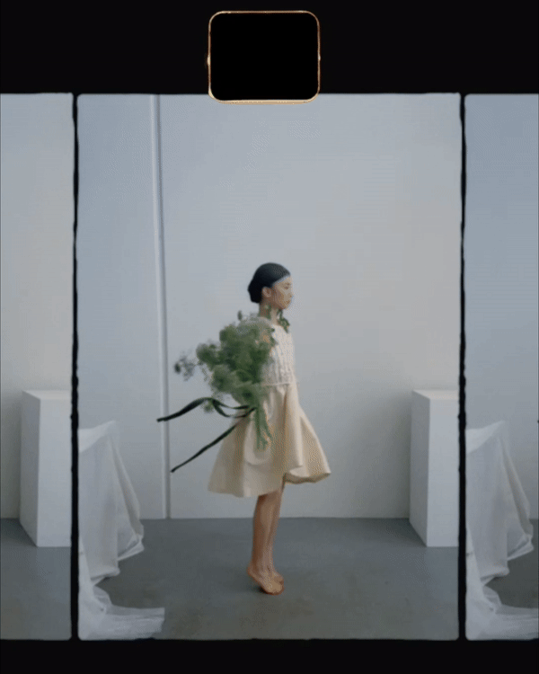 A woman standing in a minimalist white room, holding a large bouquet of flowers with green foliage, facing sideways with her eyes closed, and dressed in a light-colored, knee-length dress.”}