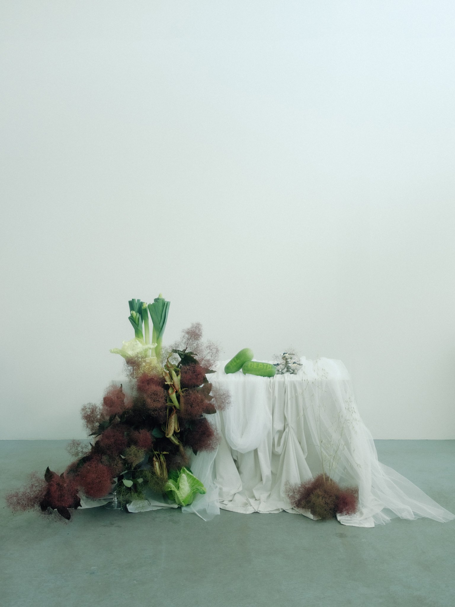 An art installation with a table draped in white fabric, green and red flowers, and objects resembling green capsules and small silver figurines, set against a plain white background.