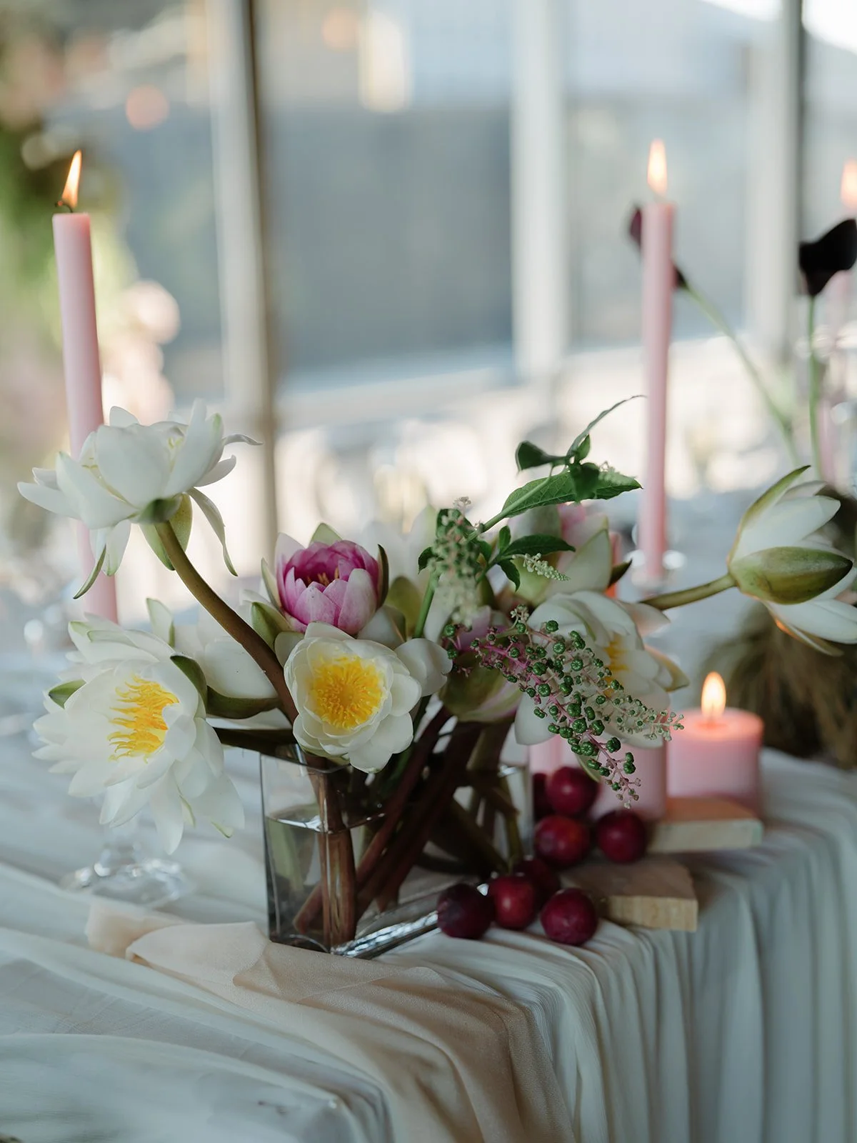 A floral centerpiece with white, pink, and yellow flowers, surrounded by pink candles, red grapes, and white candles on a table with cream tablecloth, in front of a window with blurred background.