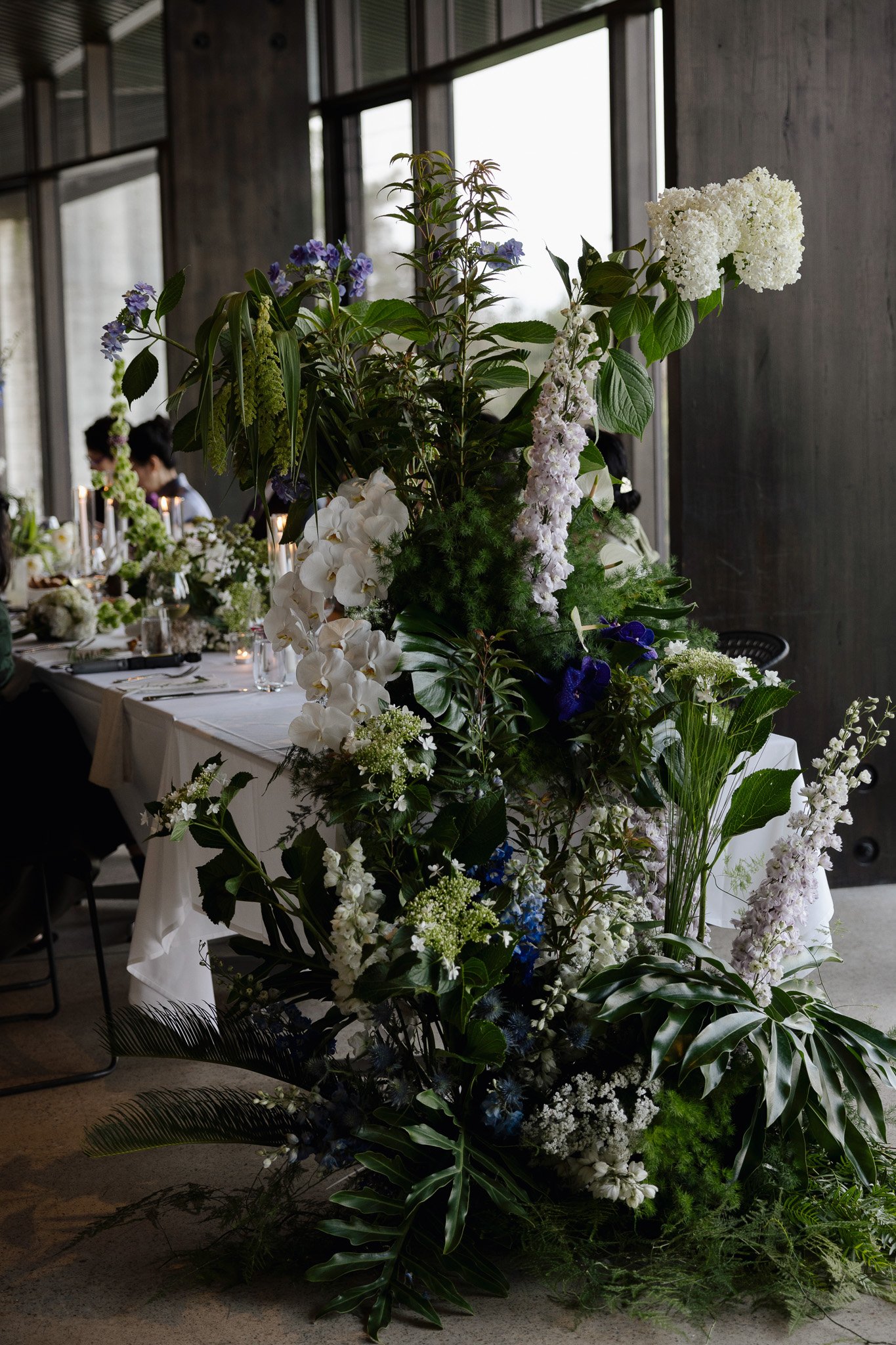Large floral arrangement with white, purple, and green flowers placed on a table inside a modern building with large windows and dark wooden walls.