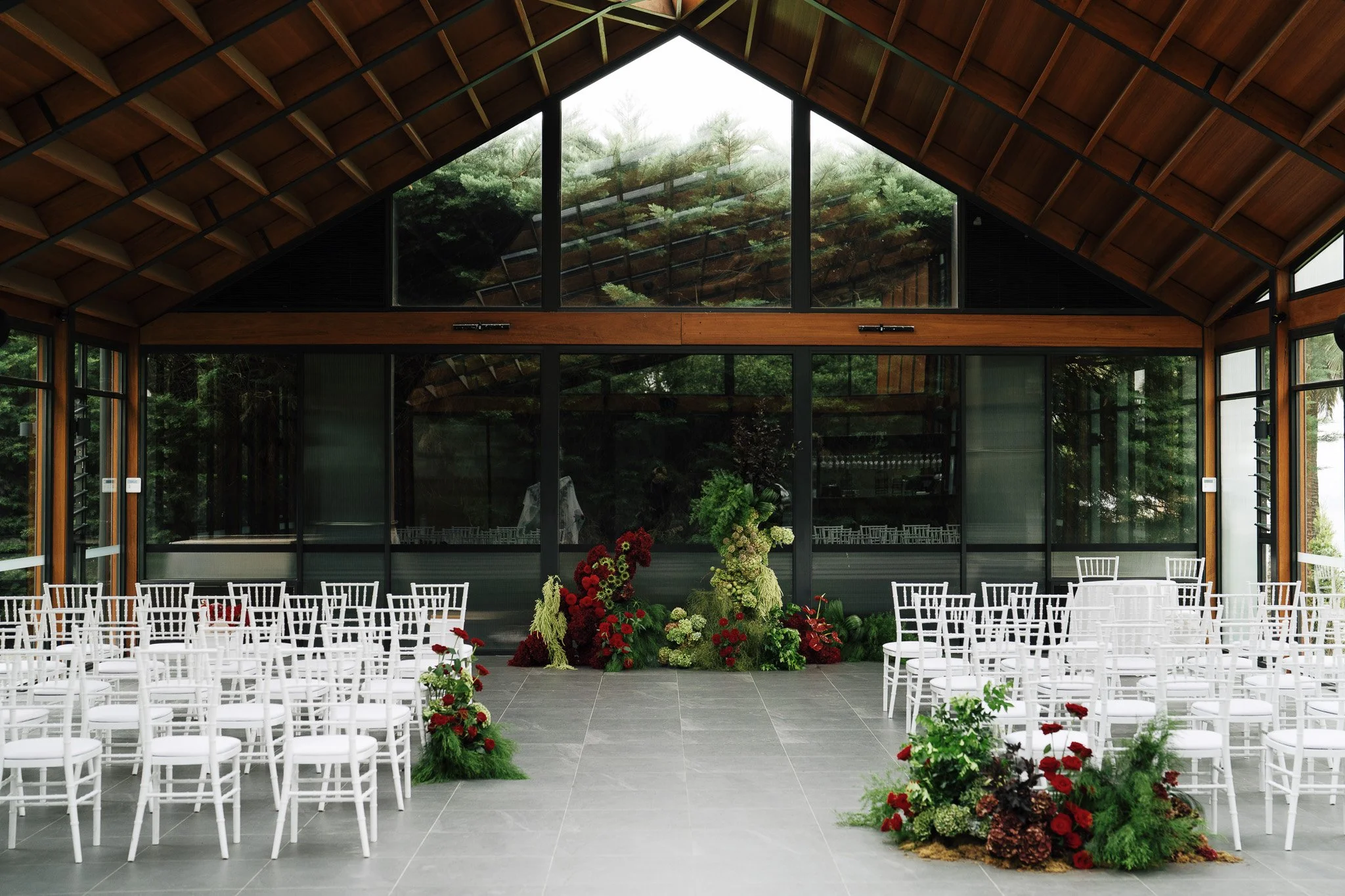 Indoor wedding or event space with white chairs arranged in rows, floral arrangements in red, white, and green, large windows, and a high wooden ceiling with a triangular glass panel showcasing trees outside.