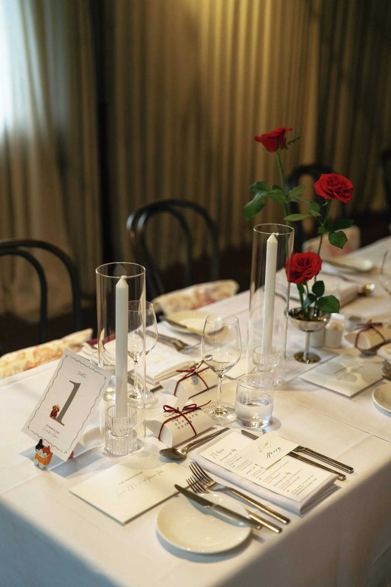 A decorated dining table set for a formal event with white tablecloth, floral centerpieces with red roses, tall glass candlesticks with white candles, glassware, silverware, and place cards.