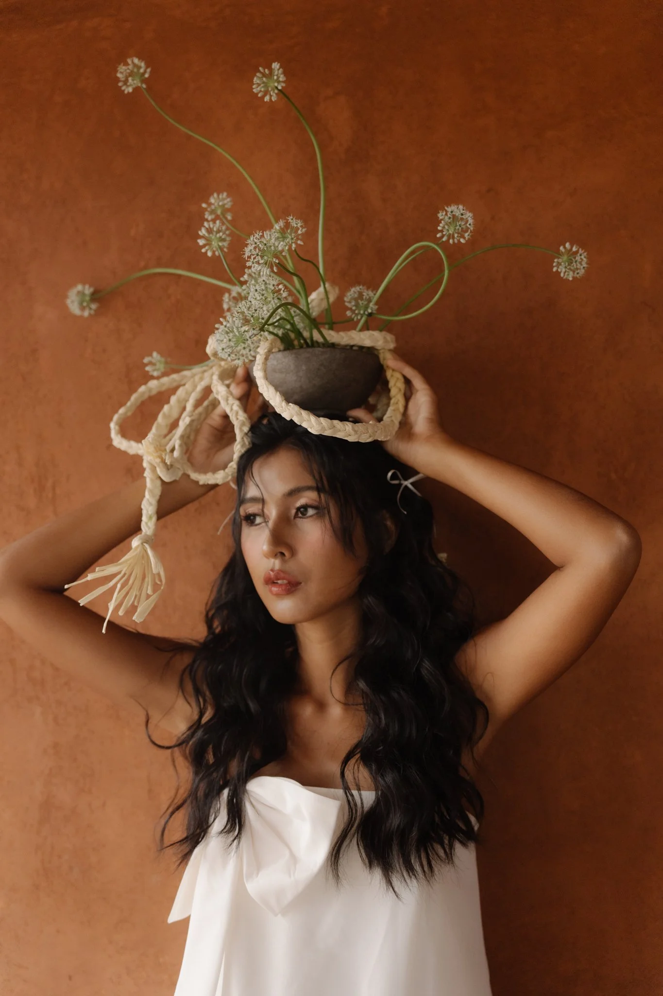 A woman with long dark hair holding a plant with white flowers in a dark bowl on her head while wearing a white top against a brown textured background.