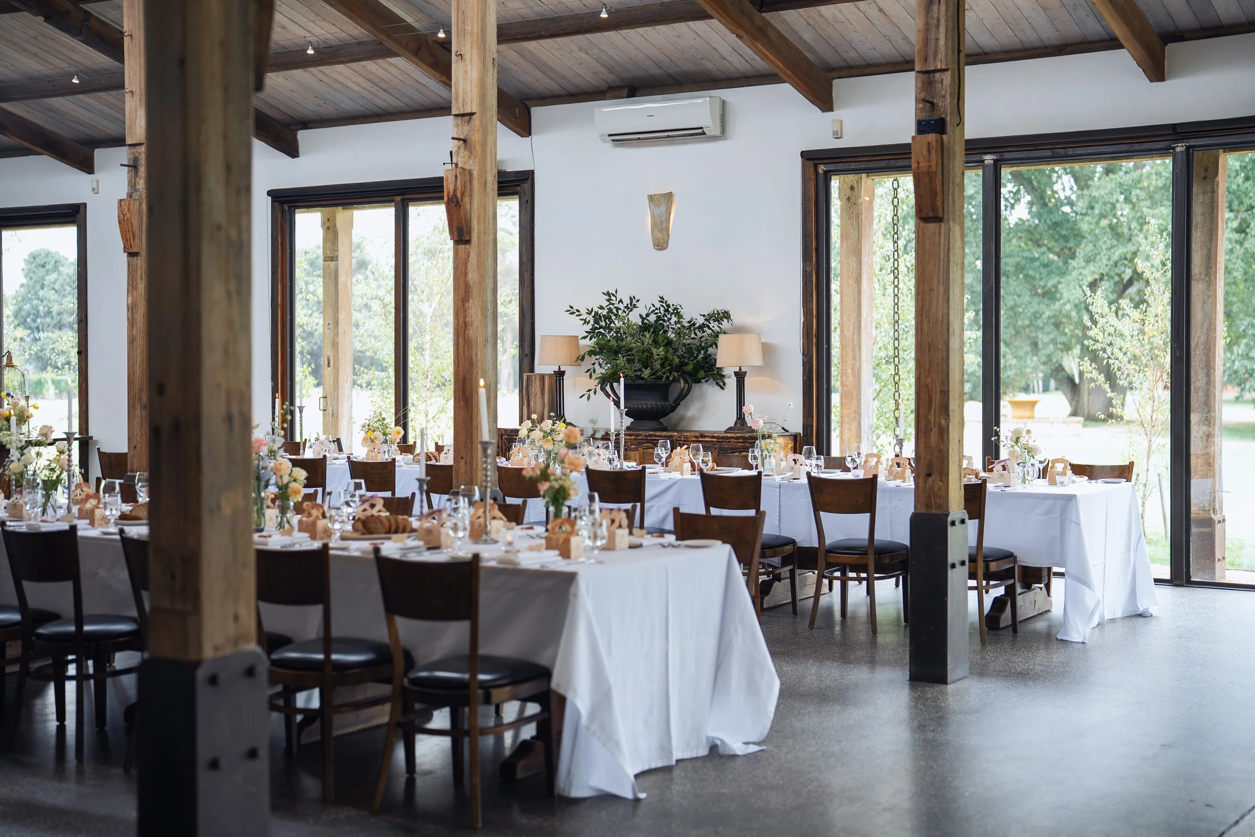 Interior of a rustic event space with long dining tables covered in white tablecloths, floral centerpieces, candles, and place settings, large floor-to-ceiling windows showing greenery outside, and wooden beams and ceiling.