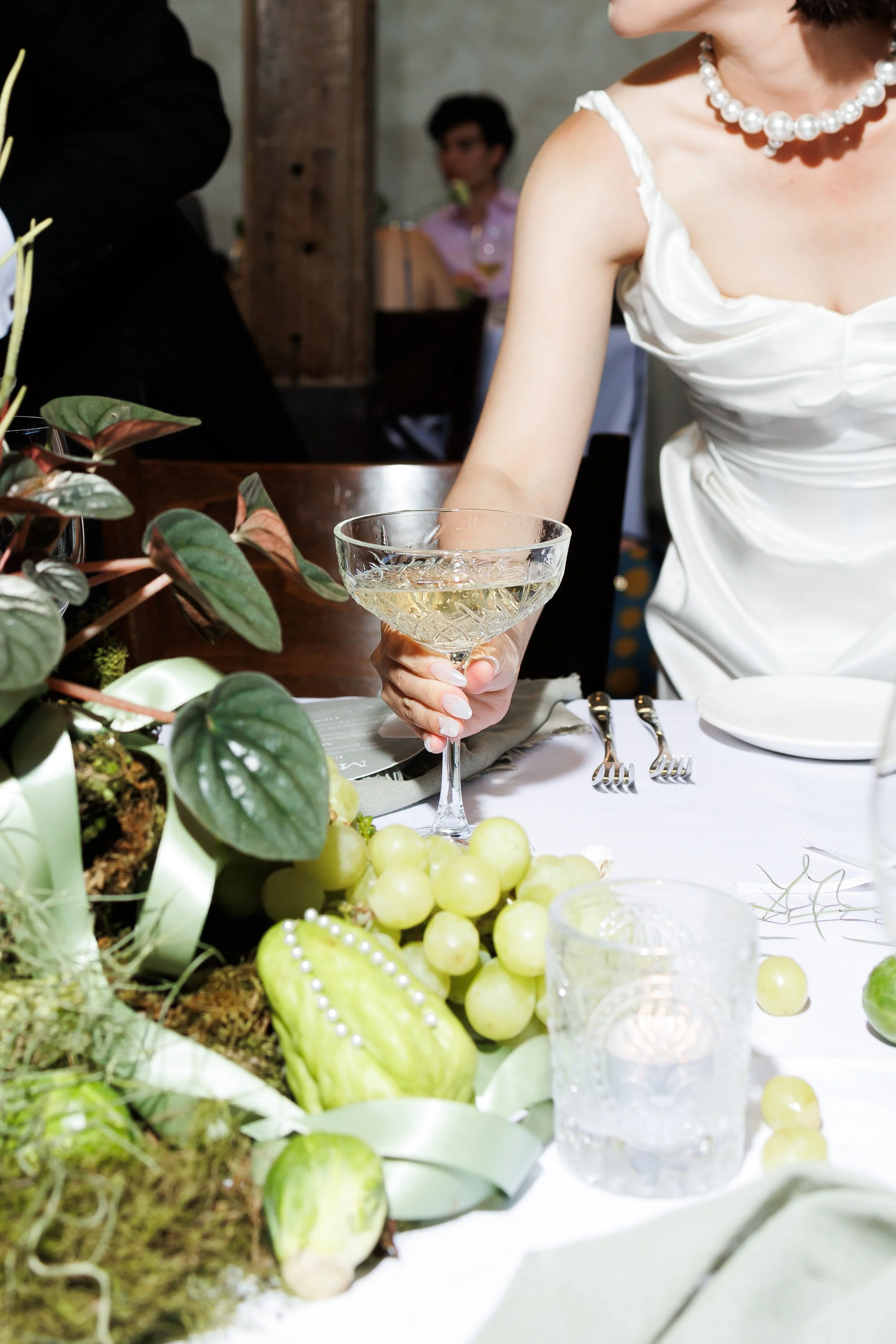 A woman in a white dress and a pearl necklace holding a champagne glass at a celebration or wedding reception. The table has grapes, green decorative gourds, and plants.